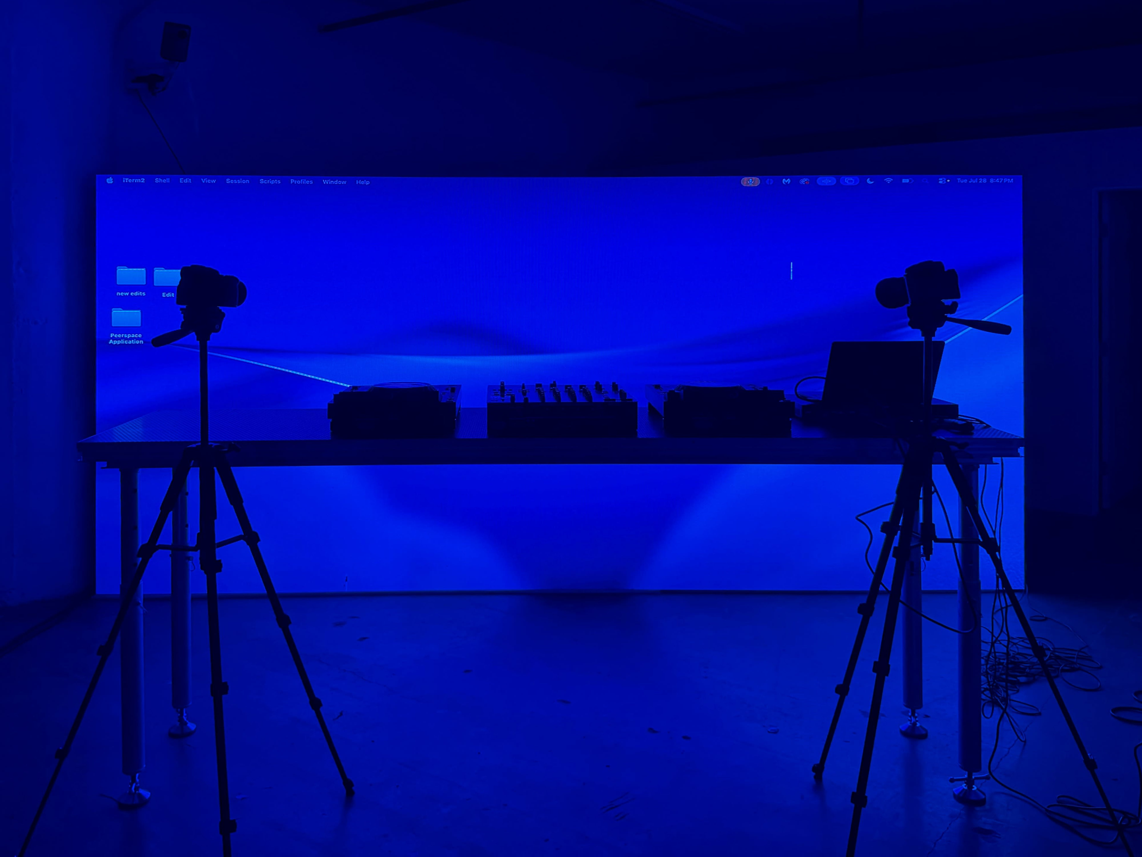 A dimly lit room features a large blue backdrop with two cameras on tripods and various audio equipment on a table.