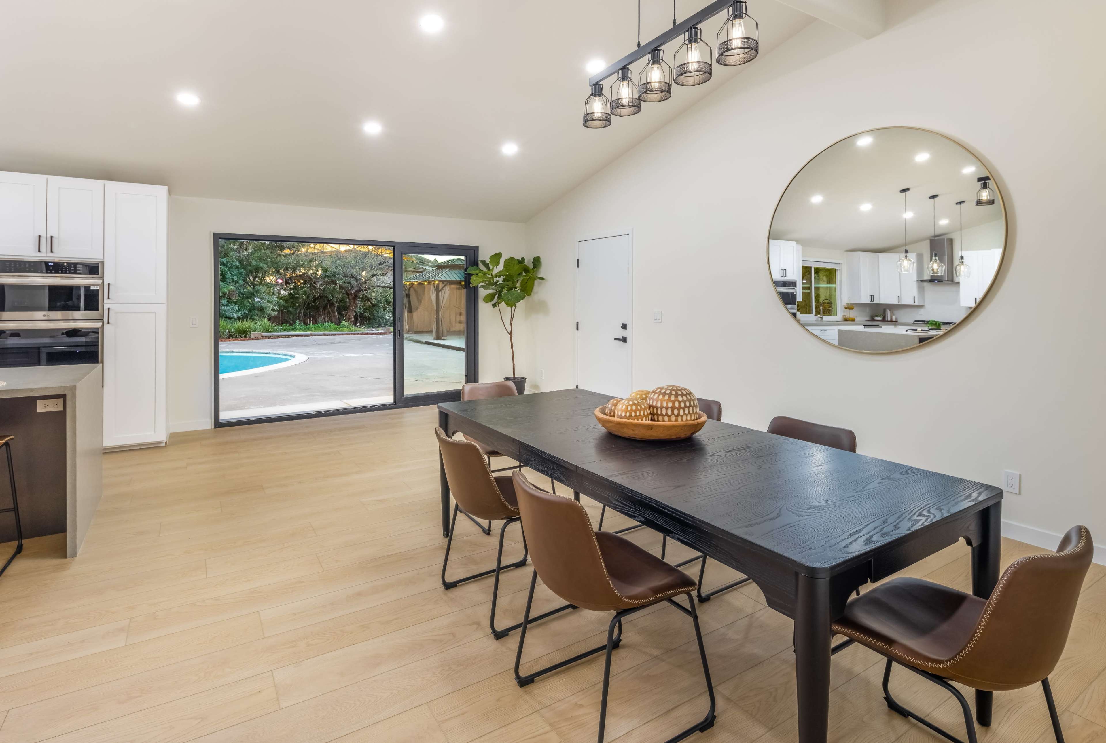 A dining area features a black wooden table surrounded by brown chairs, with large windows providing a view of a backyard pool.