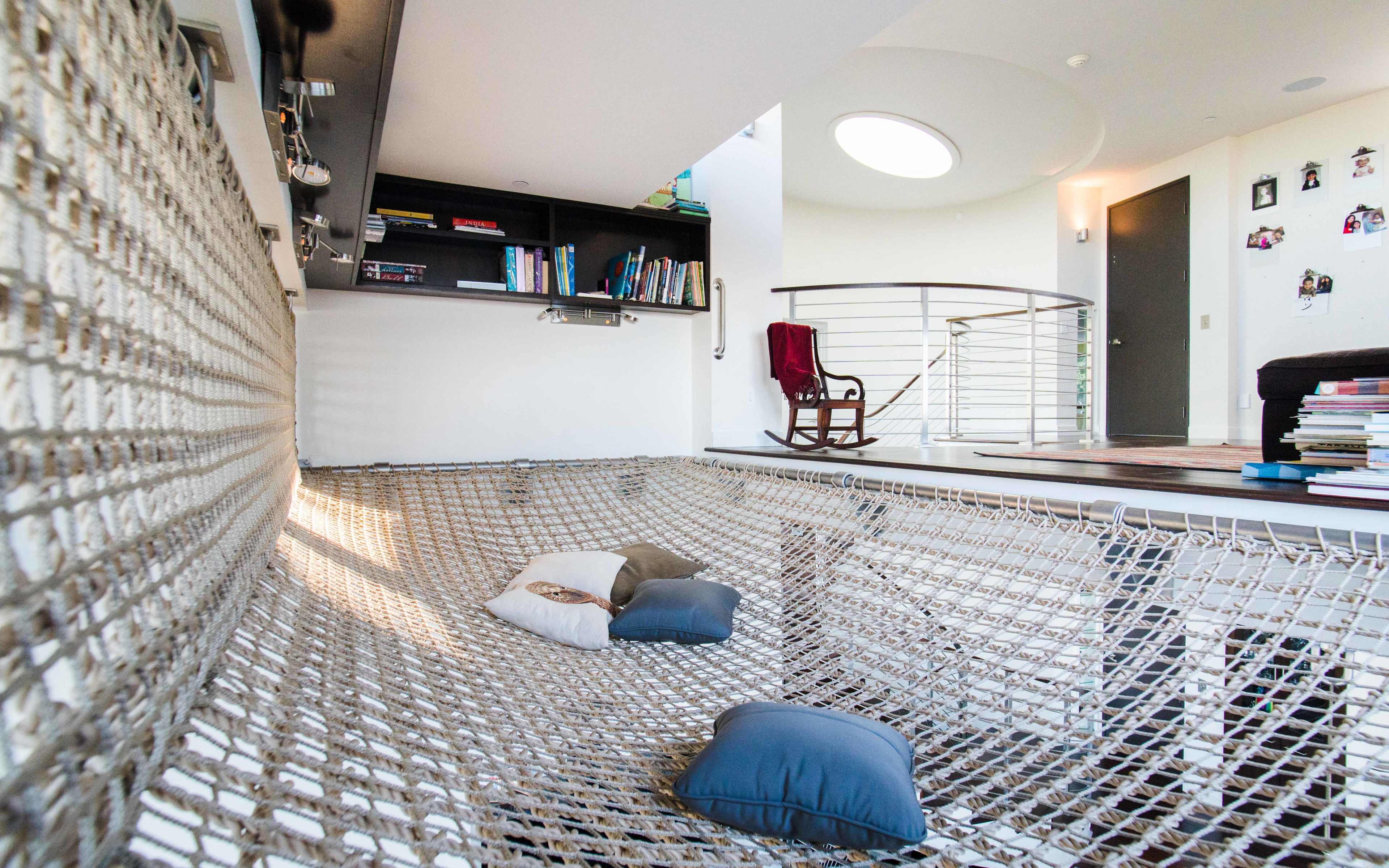 The image depicts a spacious interior with a large hammock strung between walls, surrounded by shelves of books and a staircase leading to an upper level.