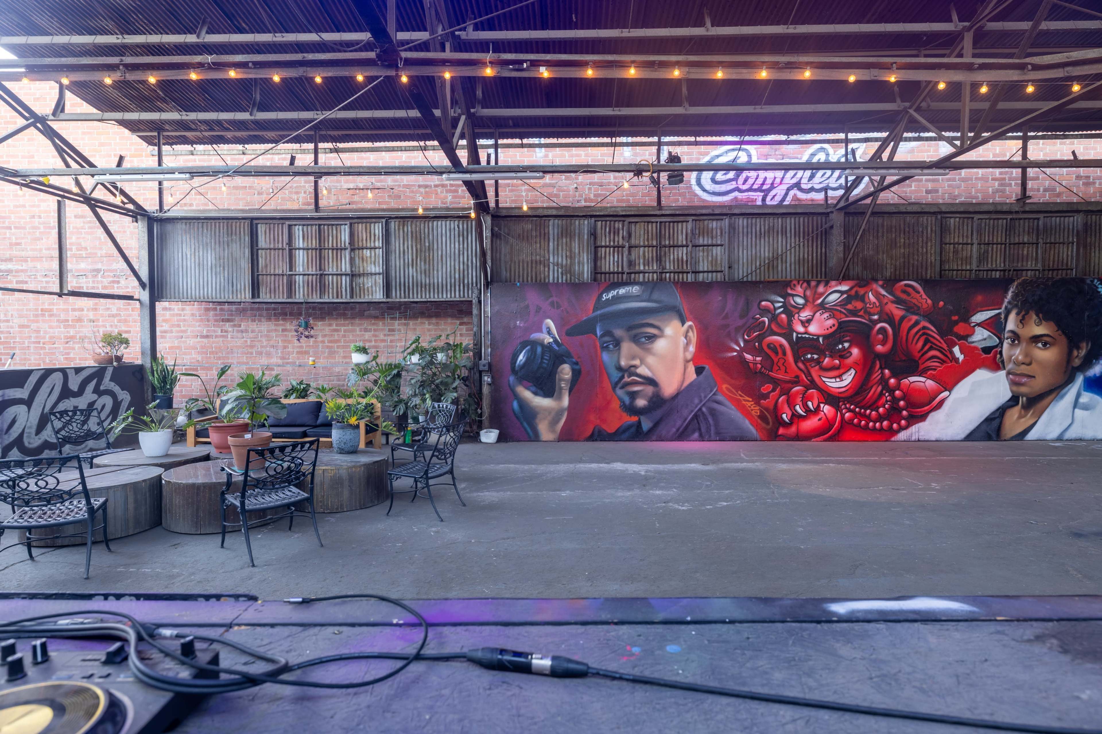 The image shows an industrial-style indoor space featuring a vibrant mural on the back wall and a seating area with tables and chairs.