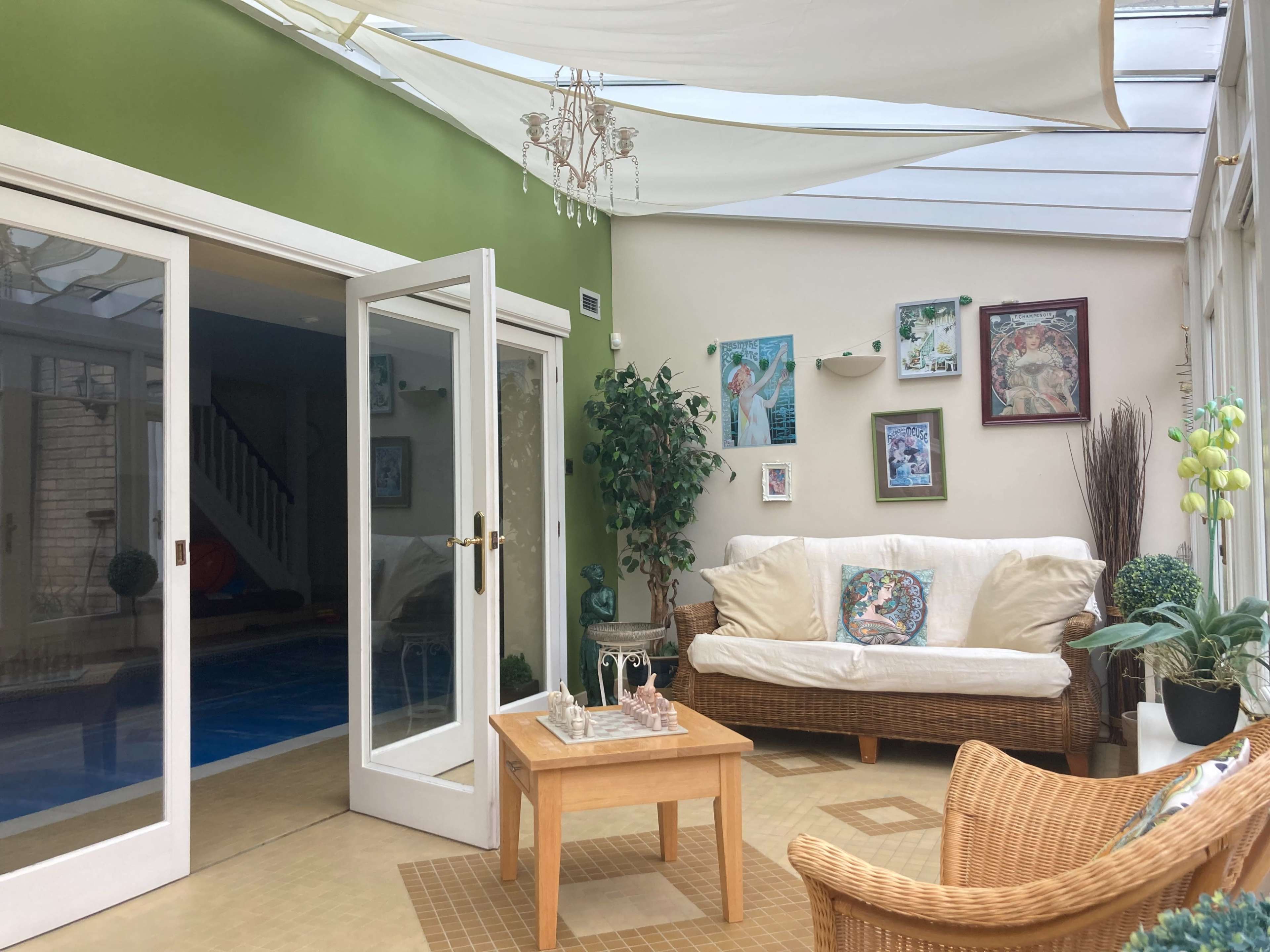 A sunlit sunroom with a white couch, a wooden coffee table, and potted plants, featuring glass walls and a clear ceiling.