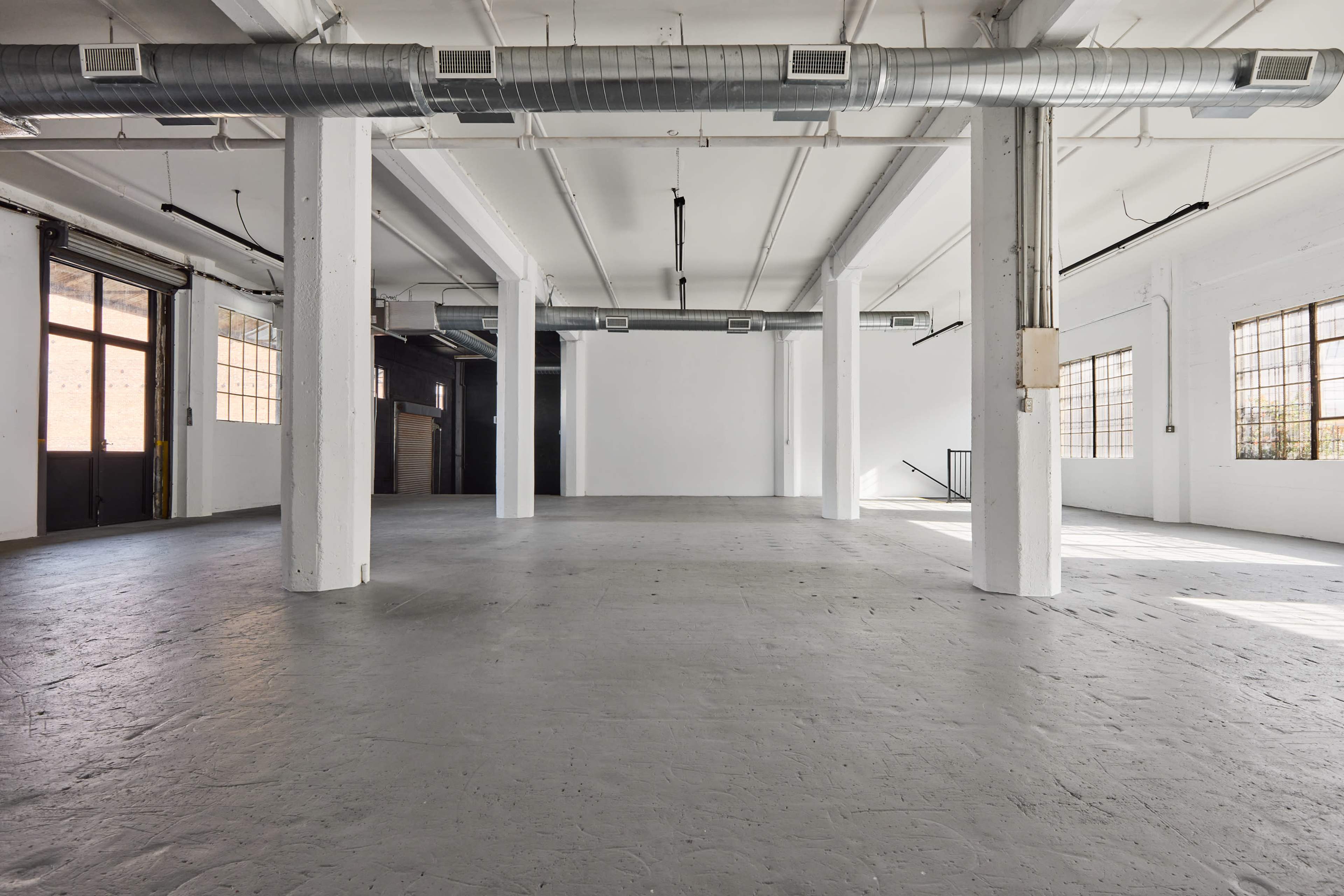 The image shows a spacious, empty industrial room with concrete flooring and large windows.