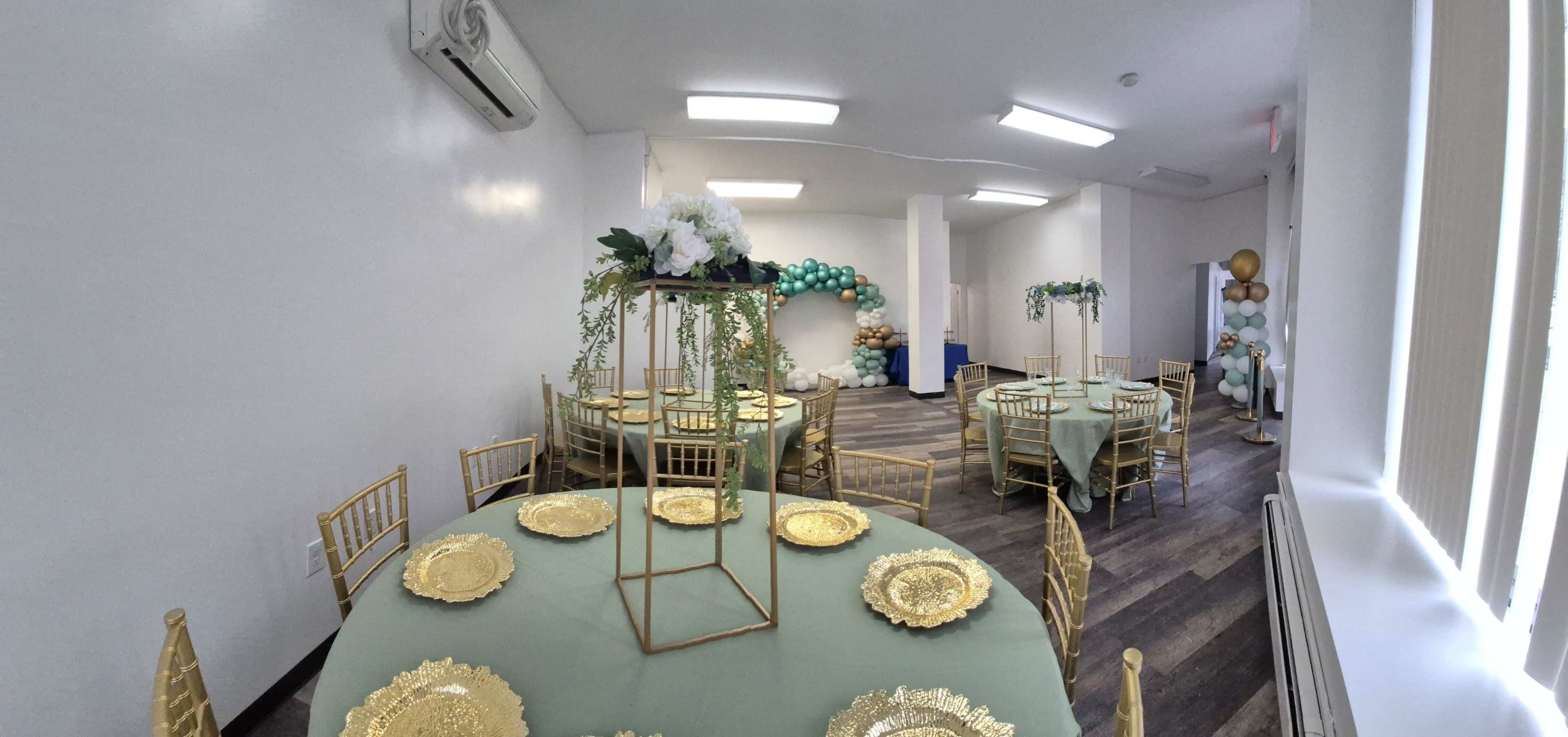 A decorated event space features round tables with green tablecloths, gold chargers, and gold chiavari chairs, adorned with floral centerpieces and balloon decorations along one wall.