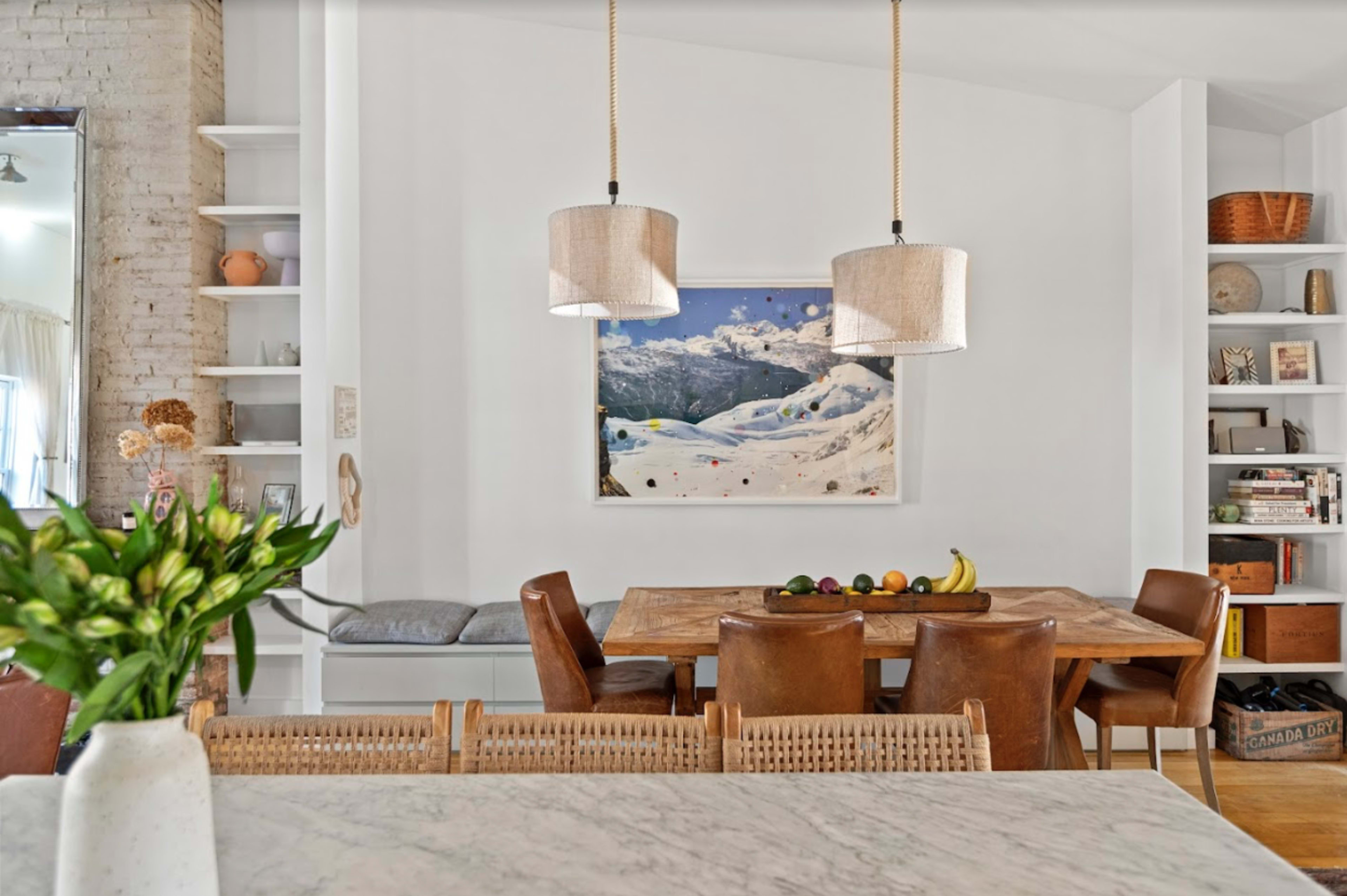 A dining area features a wooden table with various fruits, two pendant lights above, and a mountain landscape painting on the wall.