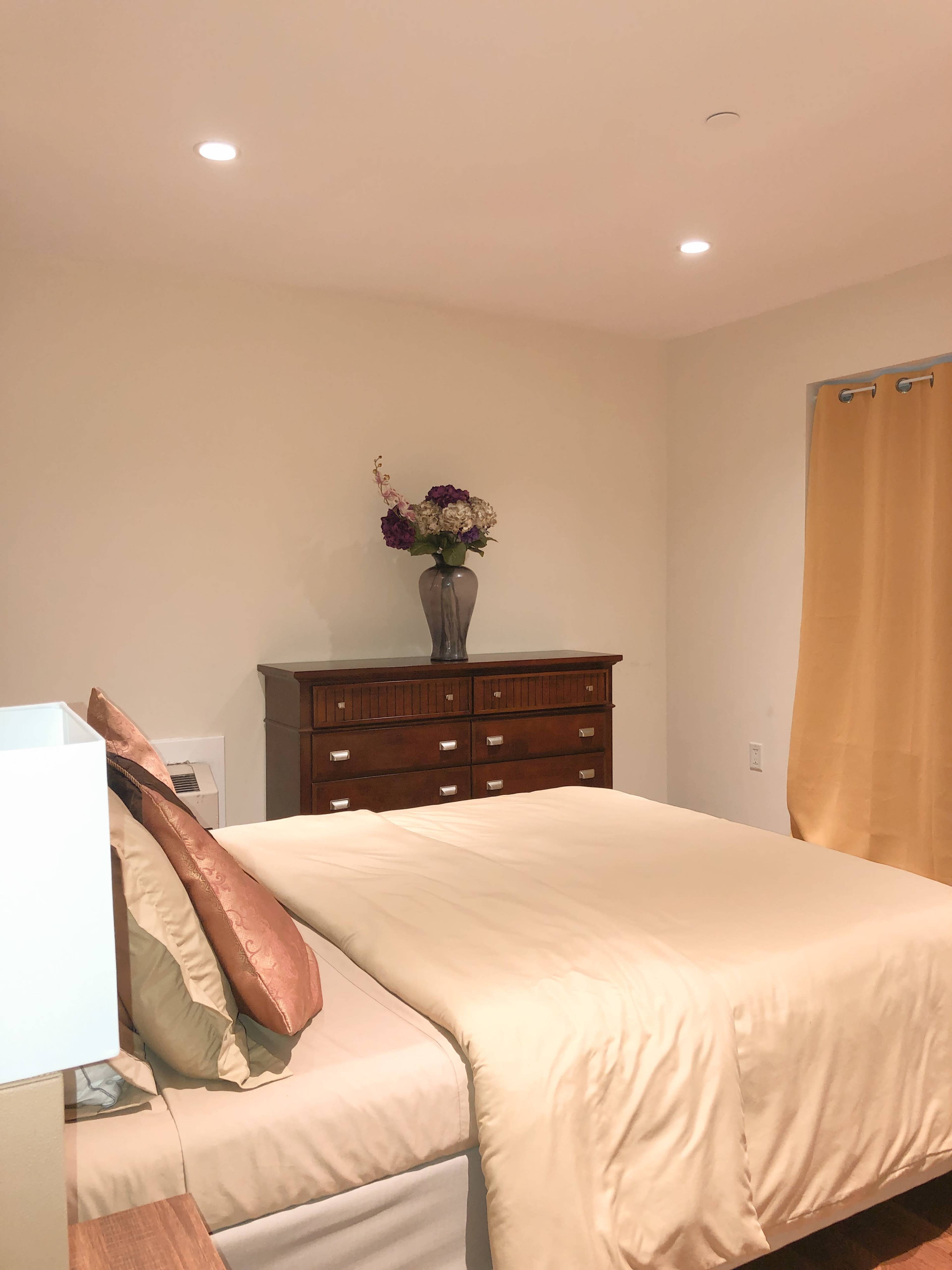 A neatly made bed with a beige comforter is positioned in a room featuring a wooden dresser and a vase of flowers on top.