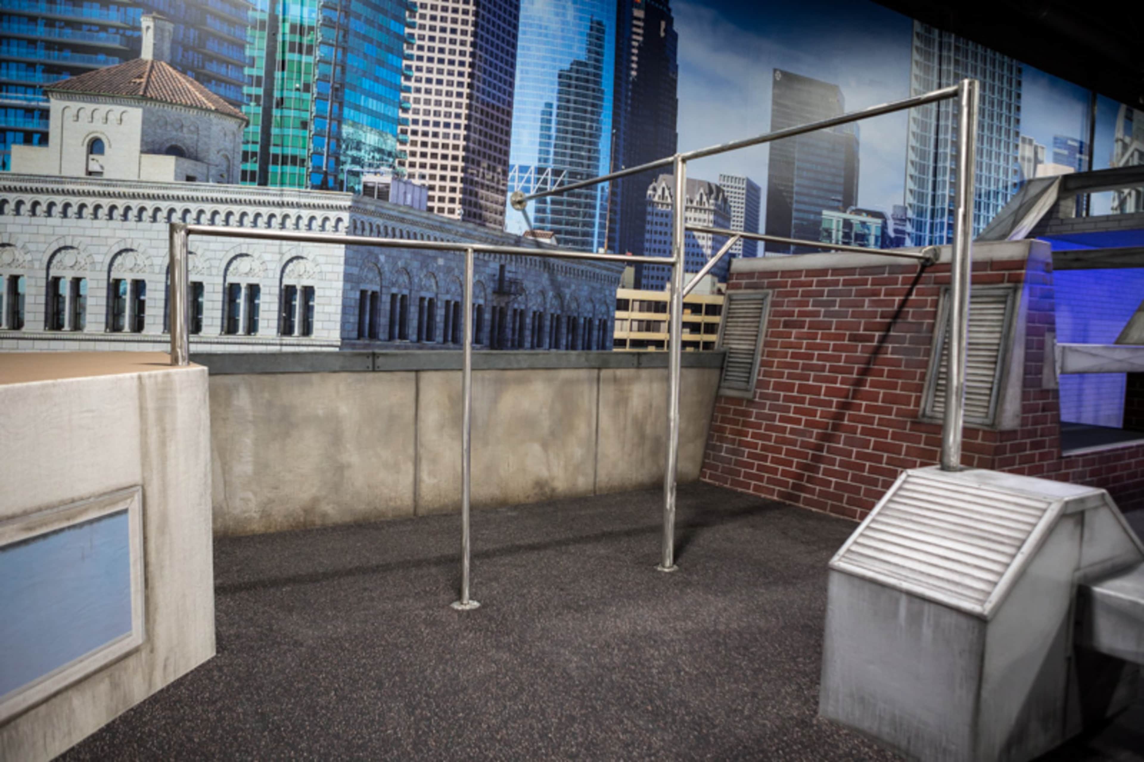 The image shows a rooftop setting with a metal framework and various urban architectural elements, including buildings and a skyline backdrop.