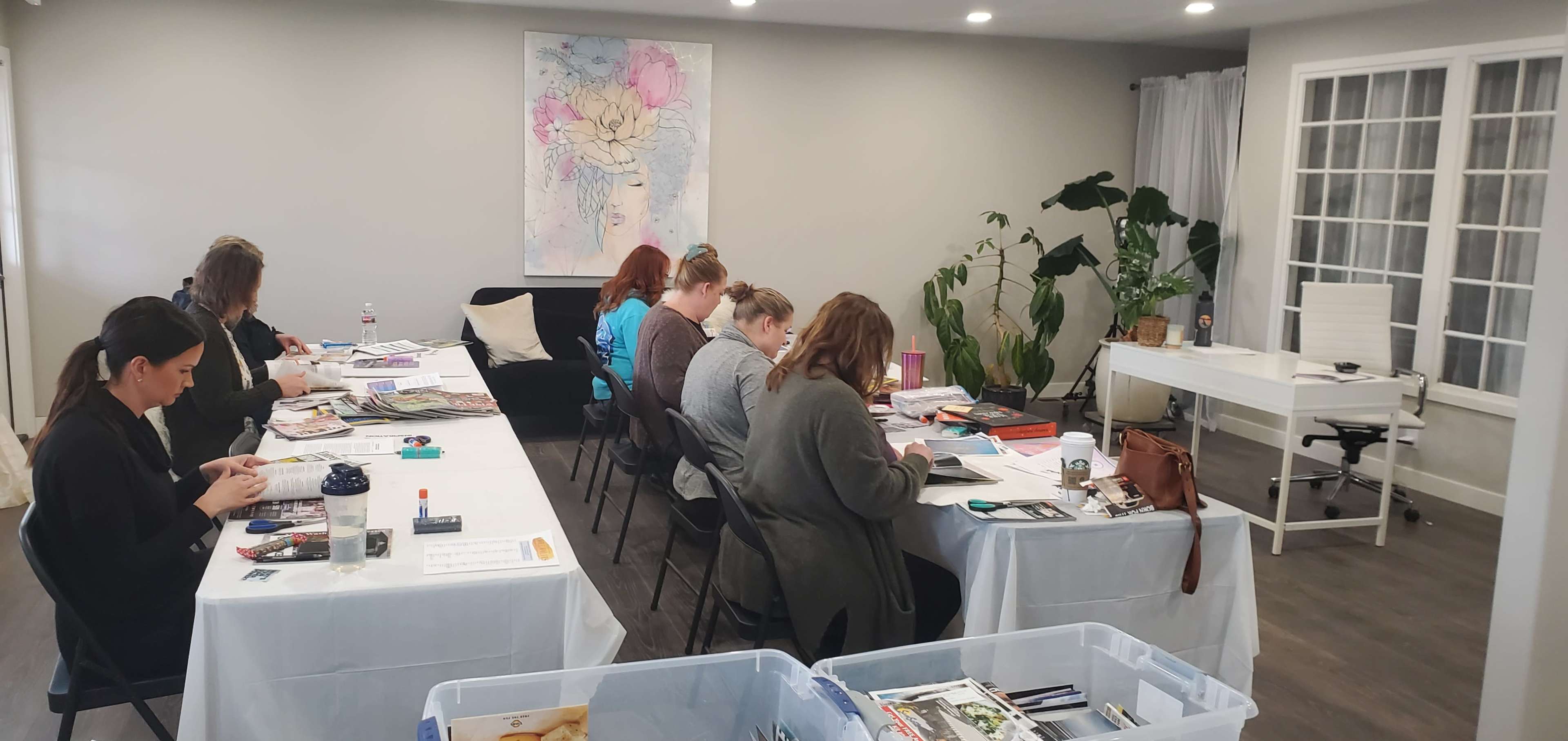 A group of seven people are seated at tables engaged in a crafting activity in a well-lit room with plants and a large artwork on the wall.