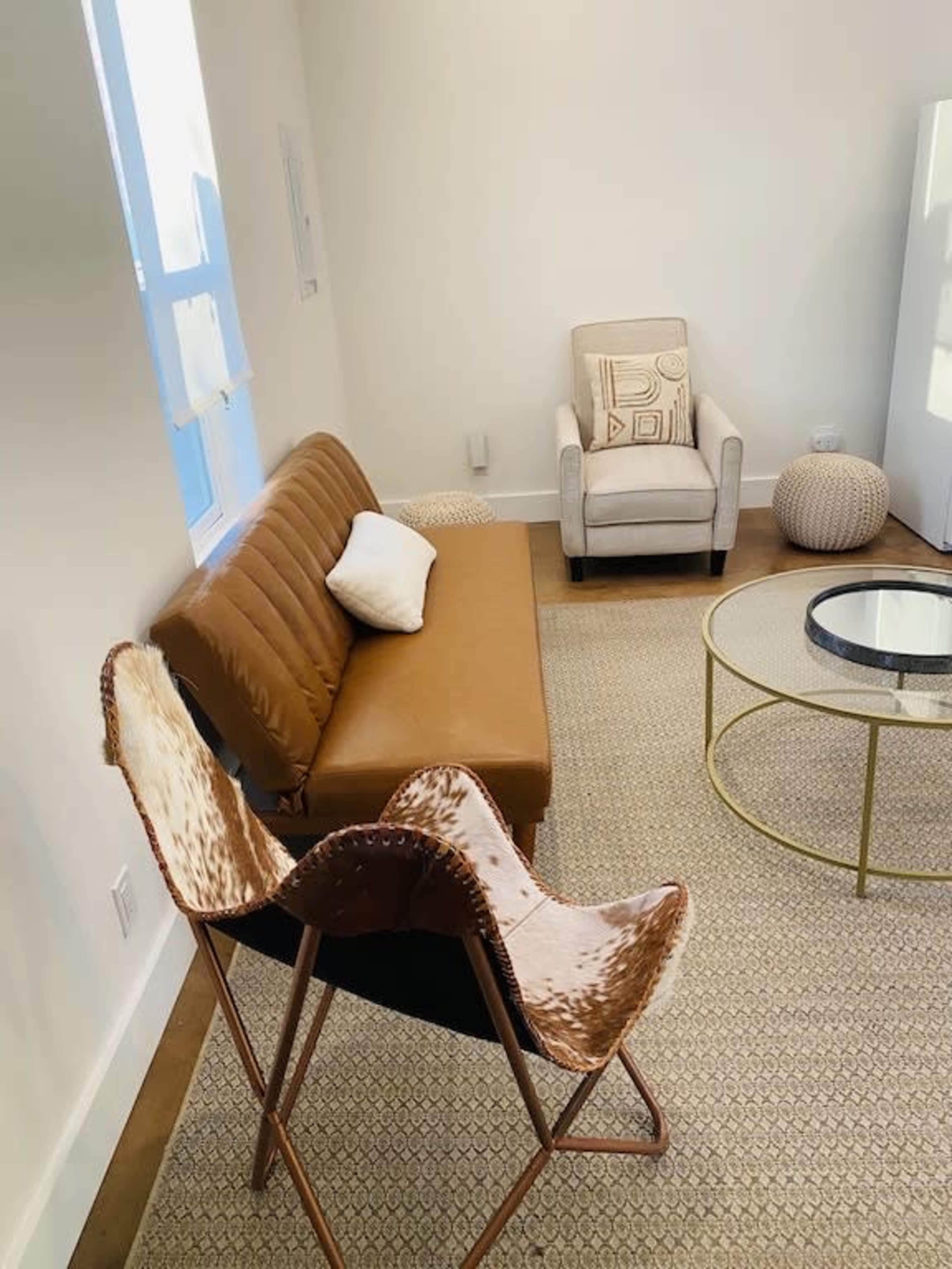 A minimalist living room featuring a brown sofa, a beige armchair, a round glass coffee table, and a patterned rug.