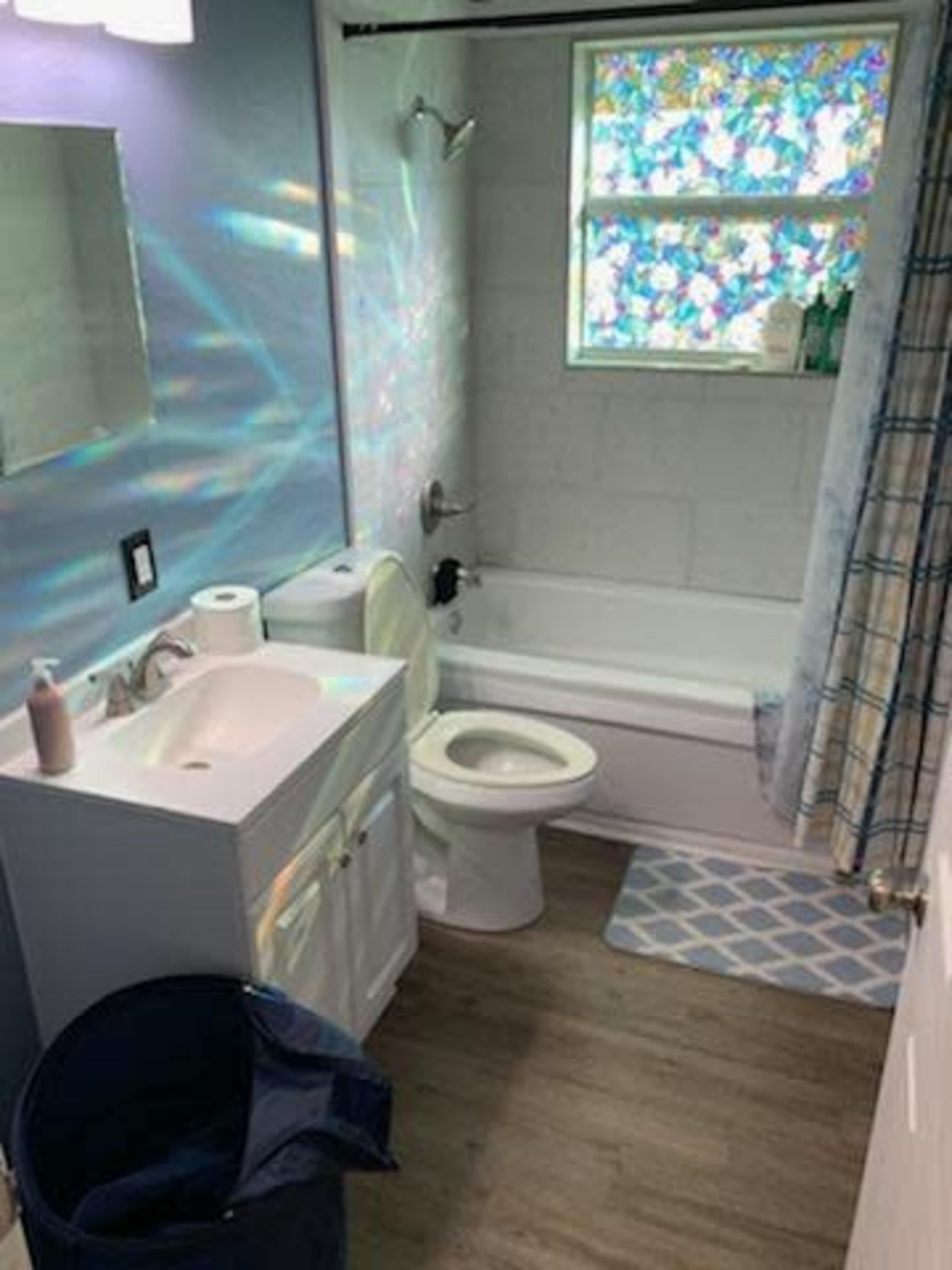 The image shows a bathroom with a bathtub, toilet, sink with a mirror, and a window featuring stained glass.