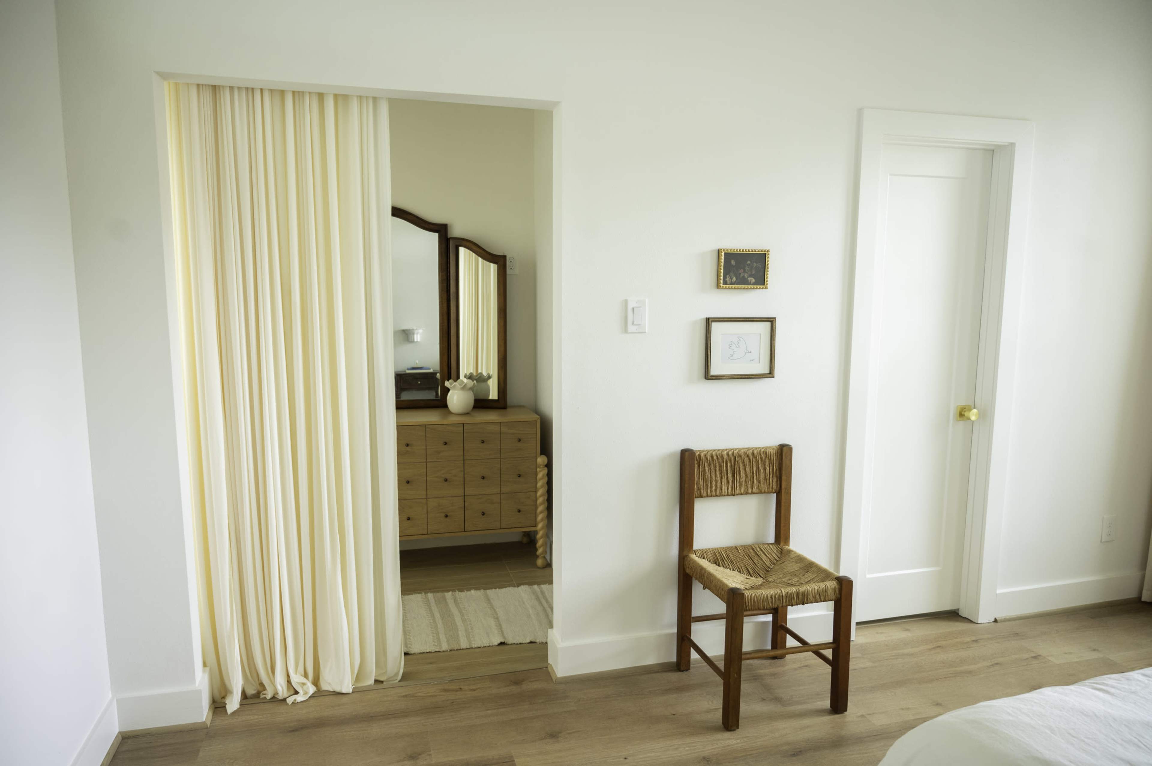 The image shows a minimalist bedroom with a wooden chair, a light curtain separating areas, a dresser with a mirror, and a door on the right.