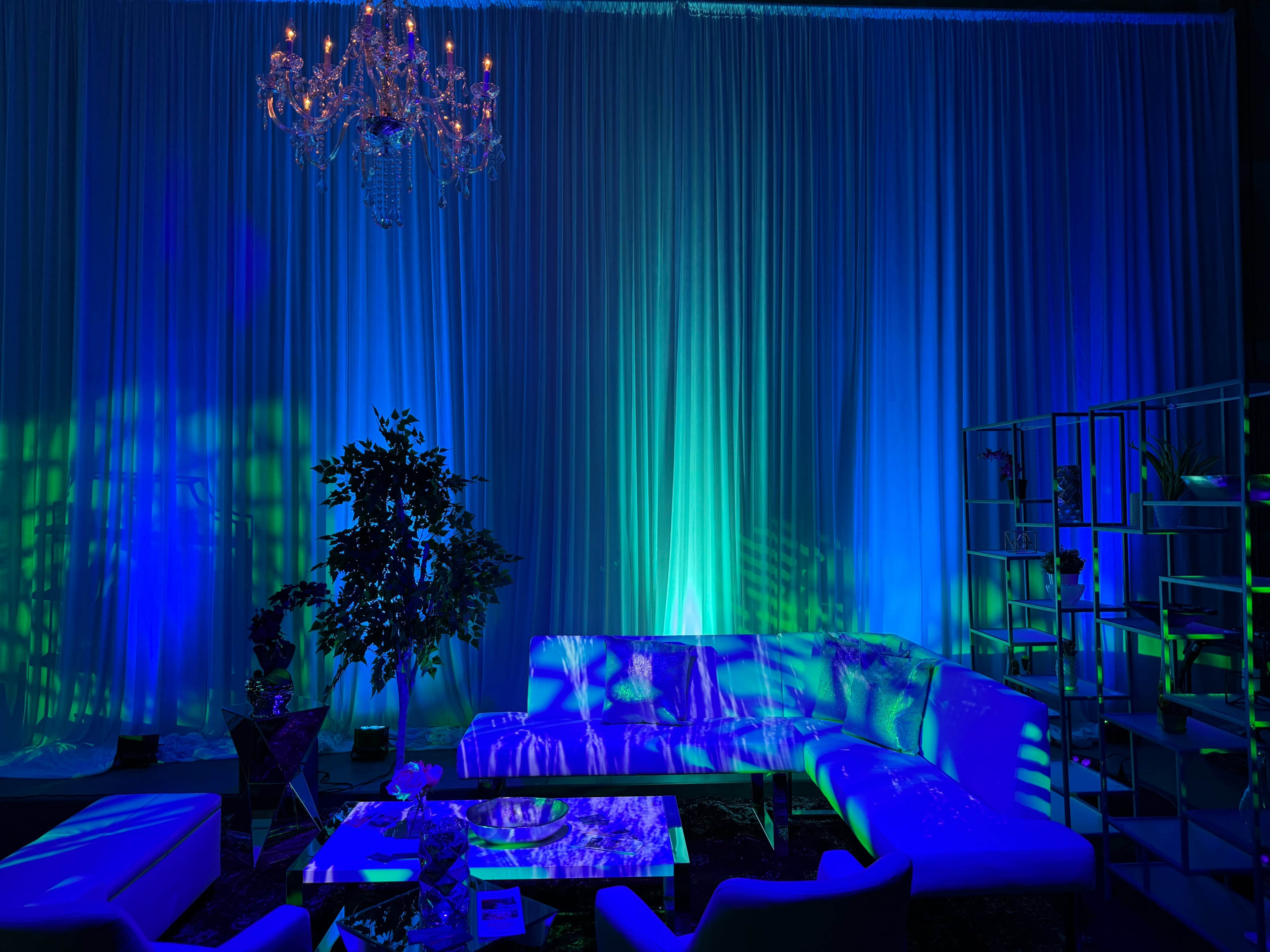 The scene features a modern lounge area with a white sectional couch, a glass coffee table, and a large chandelier, all illuminated by blue and green lights against a backdrop of draped fabric.