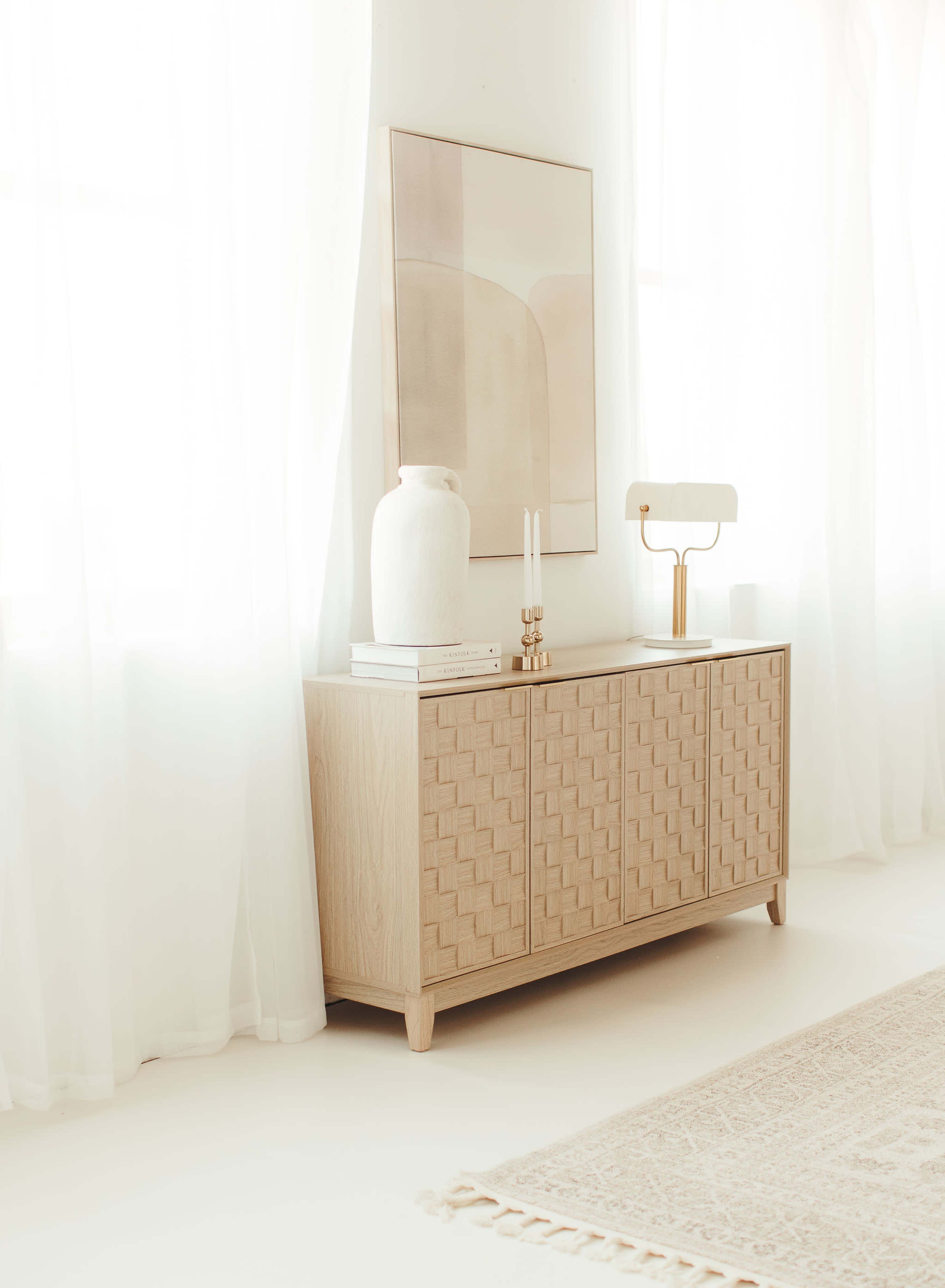 A wooden sideboard with a textured design sits against a wall, accompanied by a large vase, a lamp, and a piece of framed artwork.