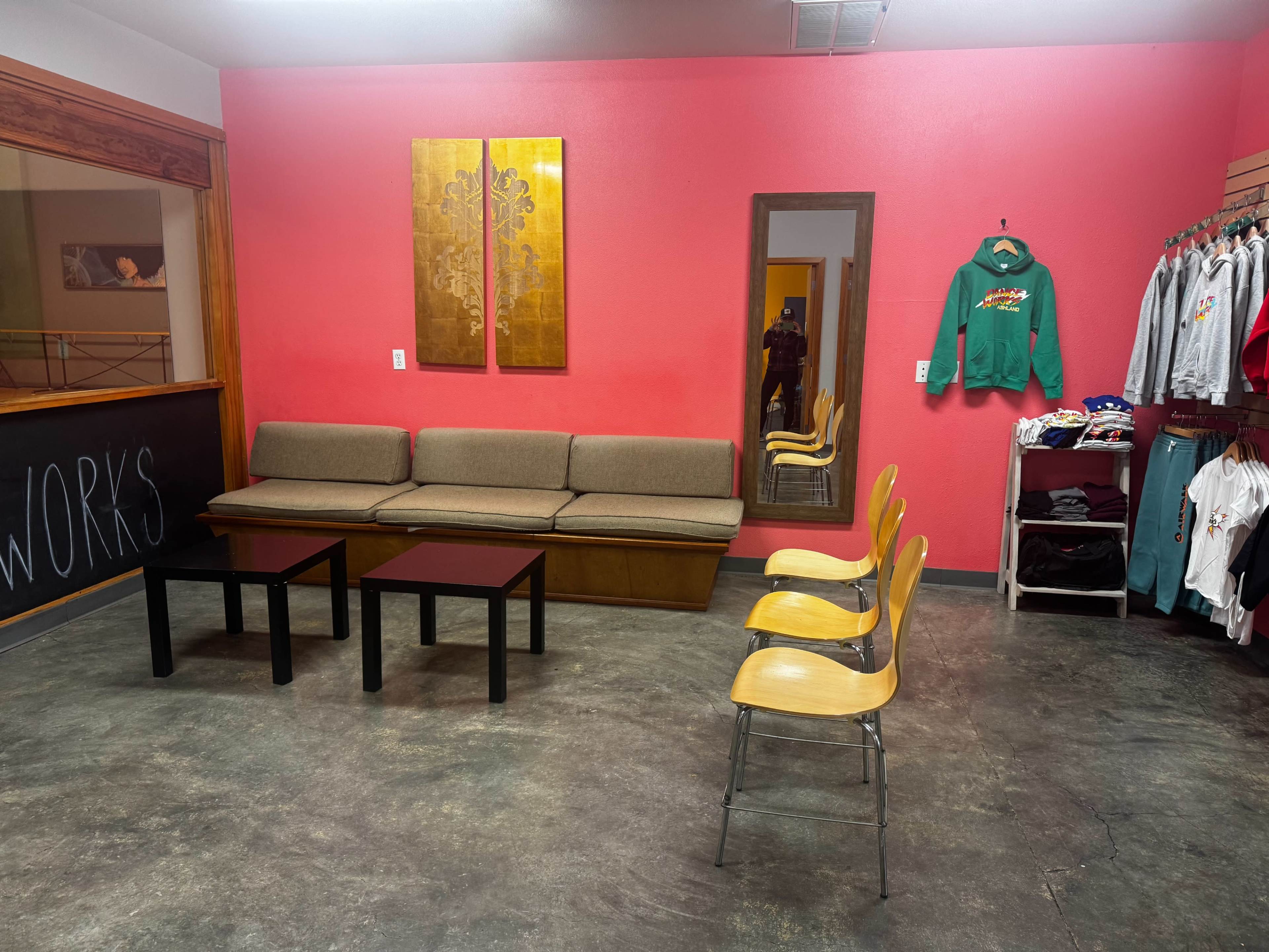 The image shows a small room with a pink wall, a brown couch, two black tables, four wooden chairs, a mirror, and a clothing rack displaying various garments.
