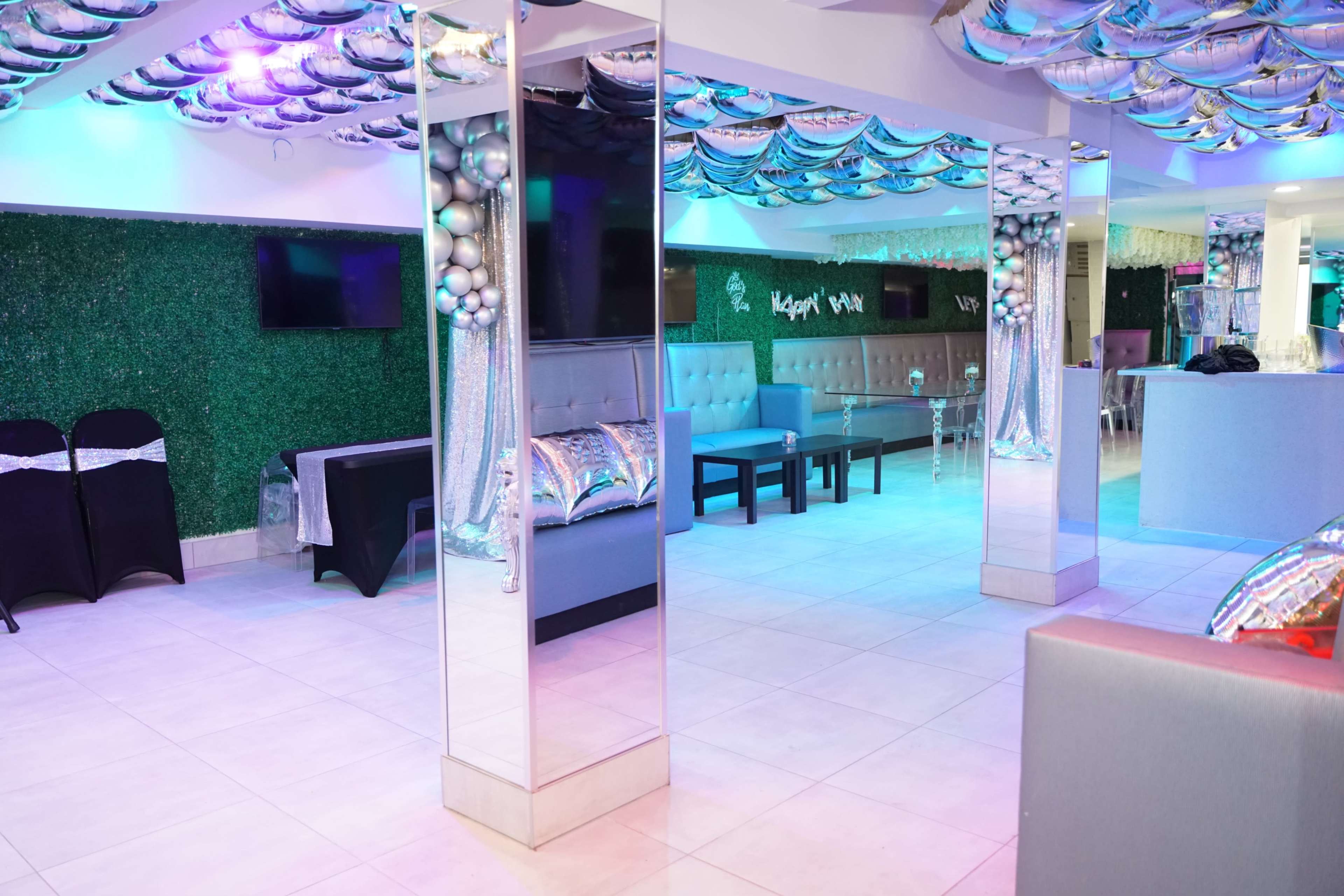 A modern event space with mirrored walls, colorful lighting, and decorative elements including silver balloons and lush green accents.