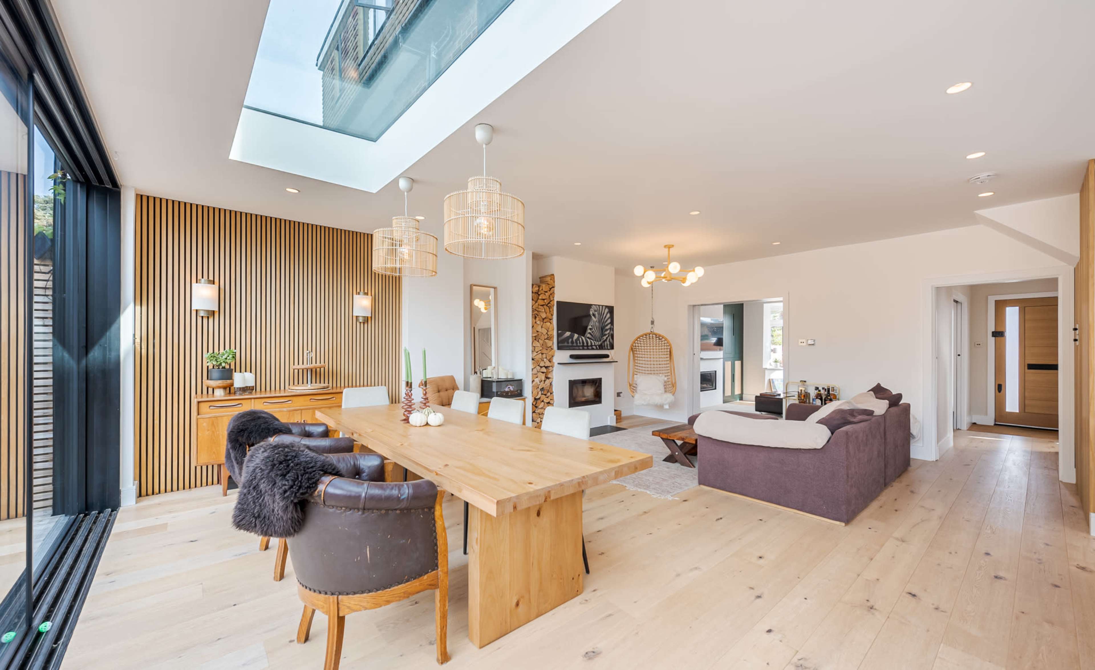 A spacious living area features a wooden dining table, upholstered chairs, and large windows with a skylight, complemented by a modern fireplace and wooden accents.