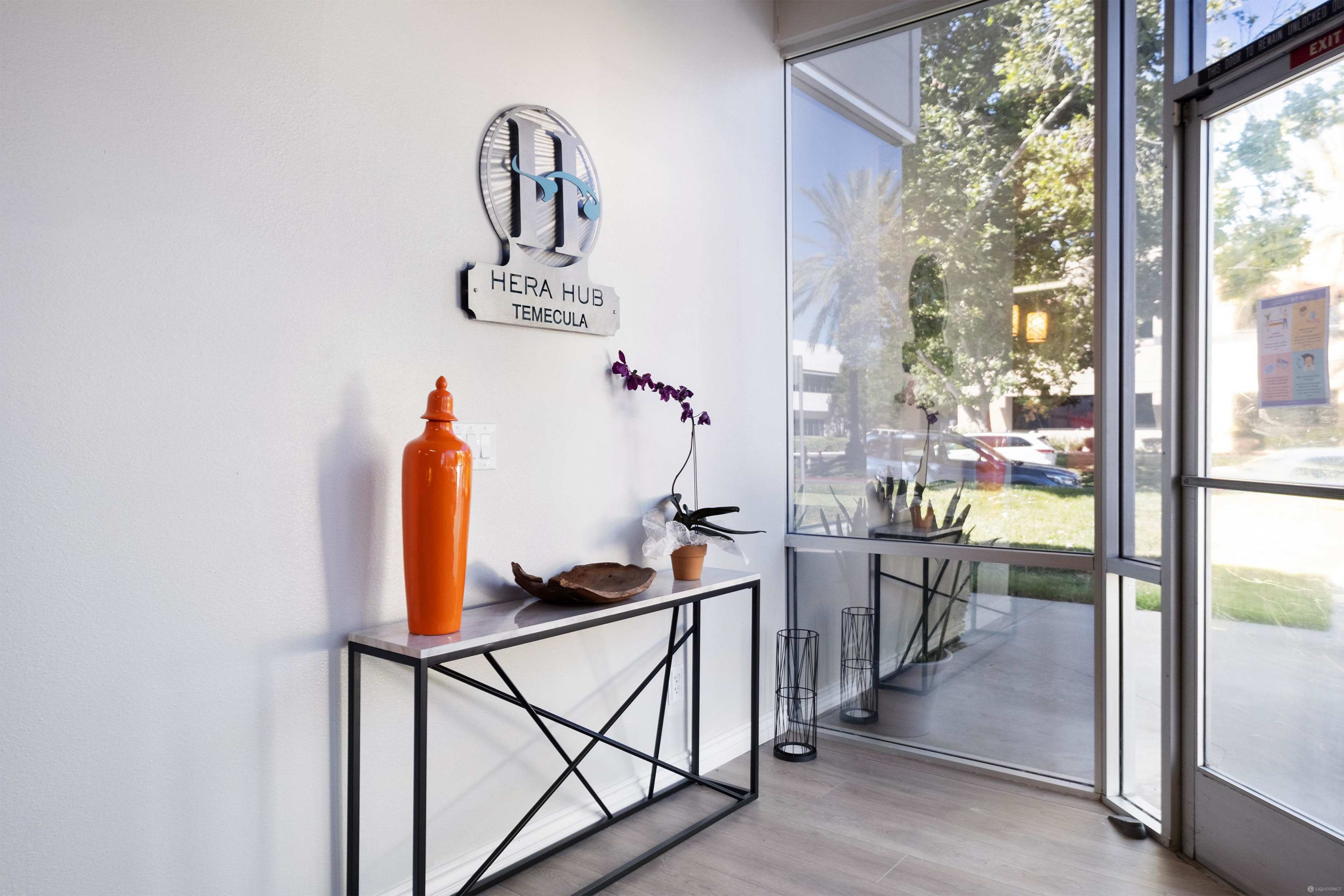 A bright entryway with a decorative table displaying an orange vase, an orchid plant, and a sign for Hera Hub Temecula on the wall.