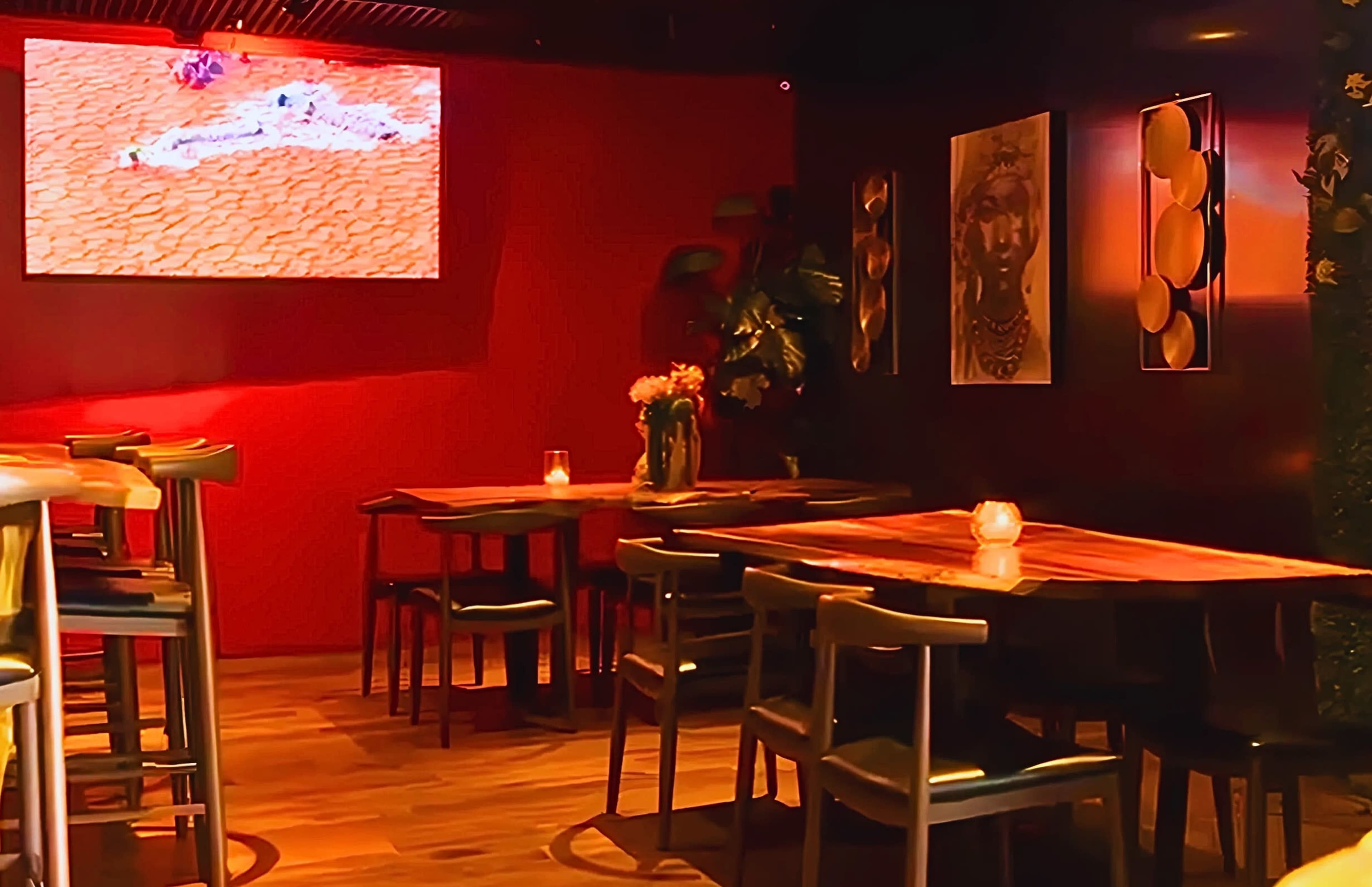 An interior scene of a dimly lit lounge featuring several wooden tables and chairs, with a large screen displaying an image on the wall in the background.
