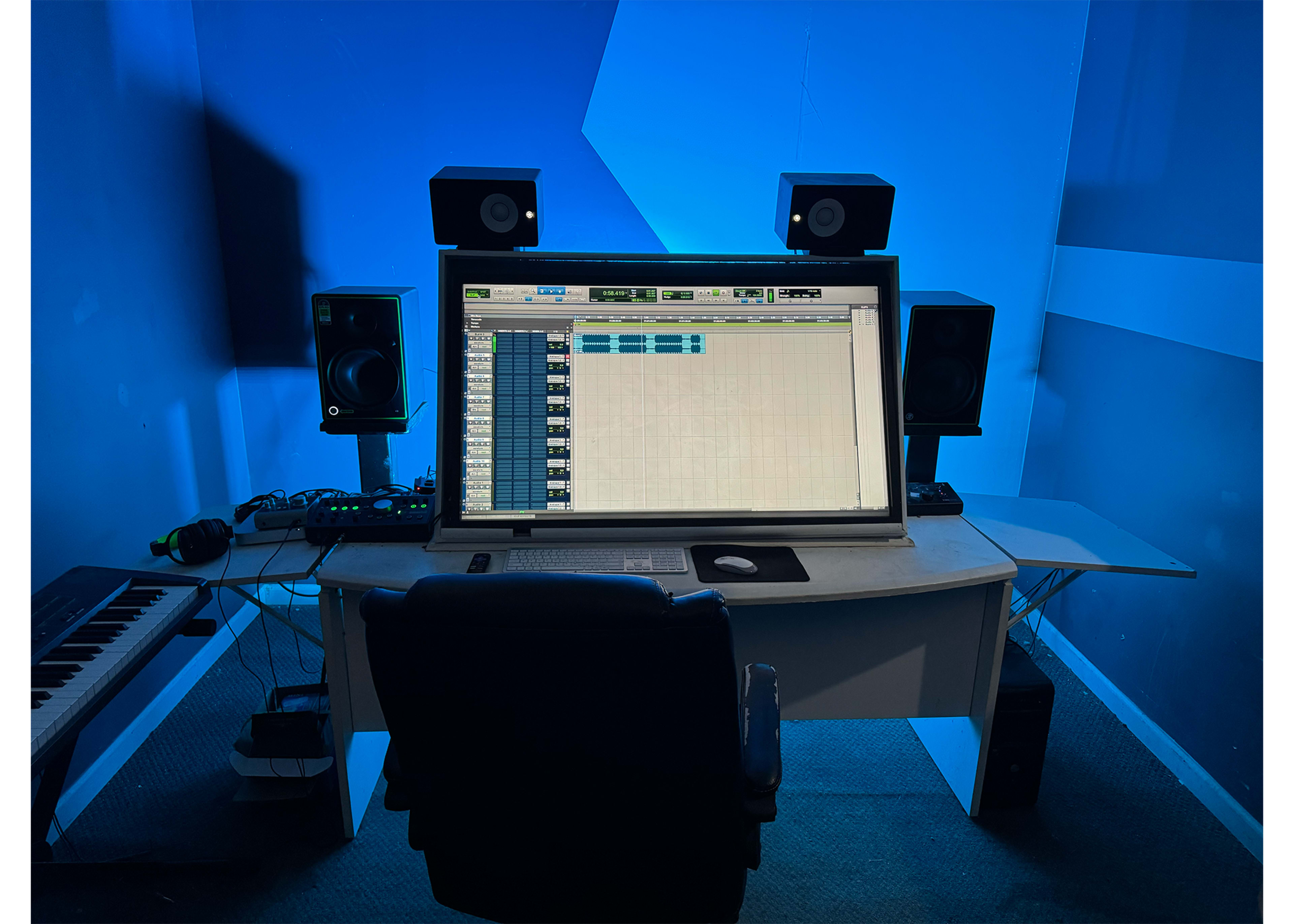 A music production studio features a large computer monitor displaying audio software, flanked by studio monitors and a keyboard in a blue-lit environment.