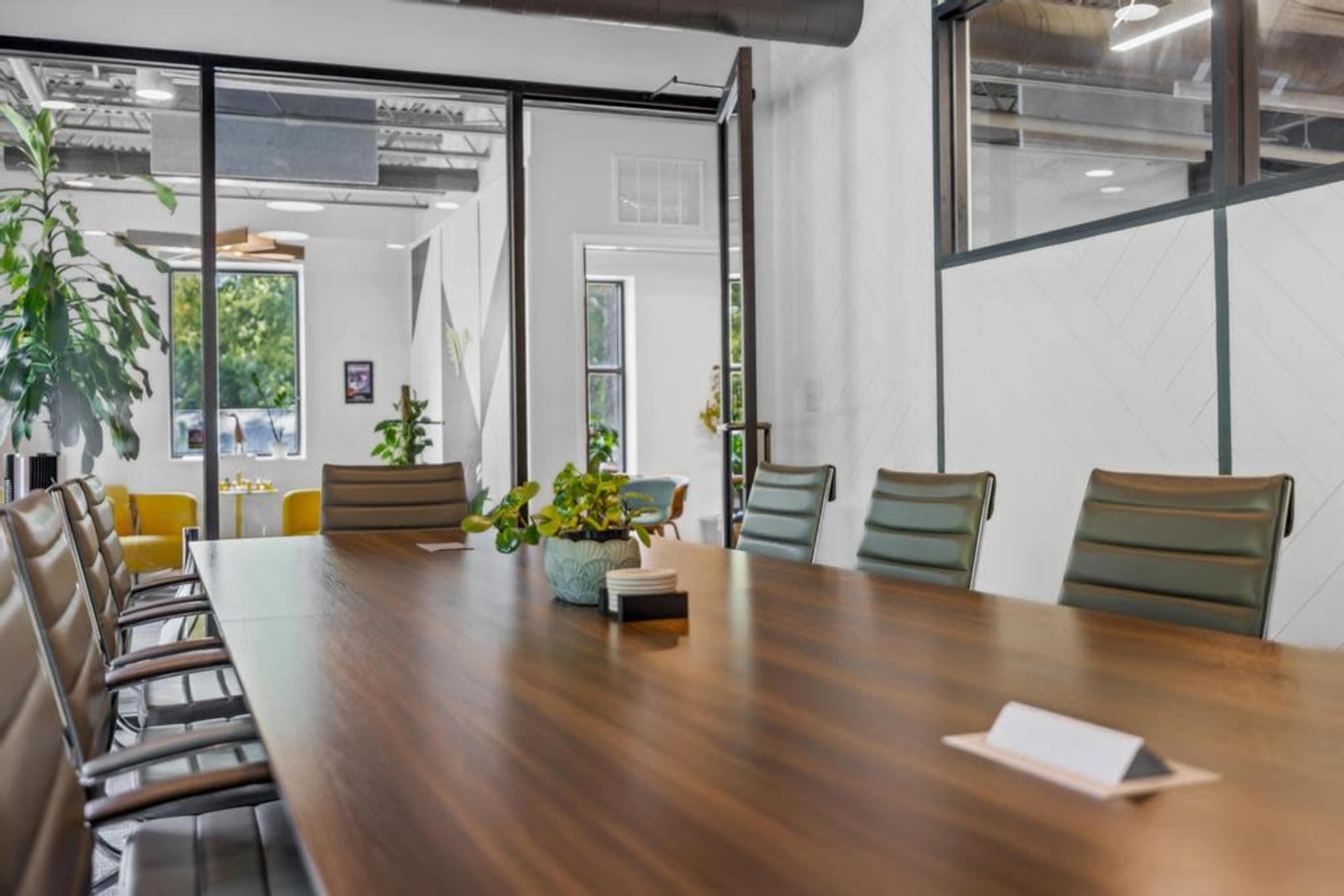 A spacious conference room features a long wooden table surrounded by leather chairs, with plants and natural light visible through large windows.