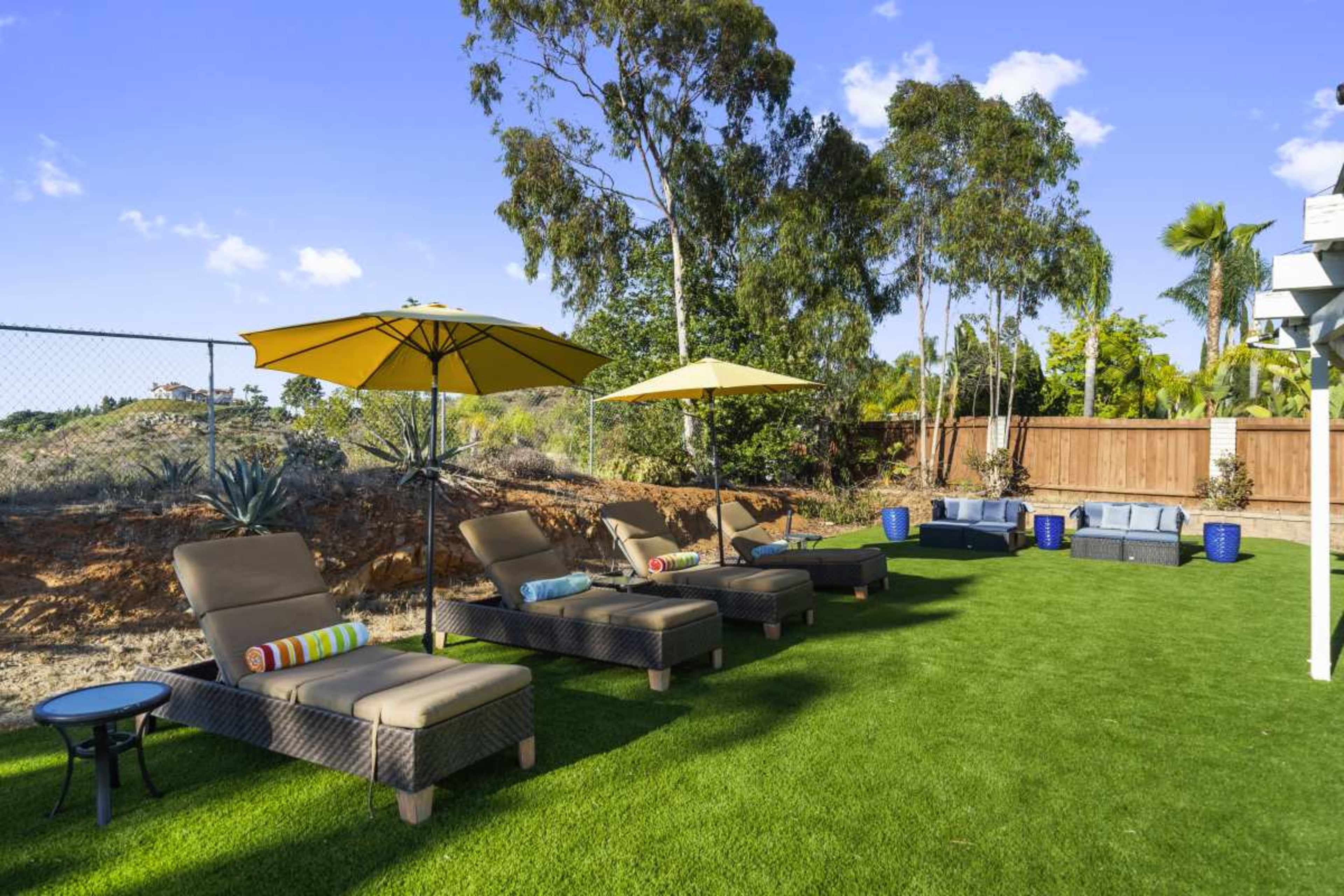 The image features a well-kept backyard with two rows of lounge chairs adorned with yellow umbrellas, set on green grass along a wooden fence.