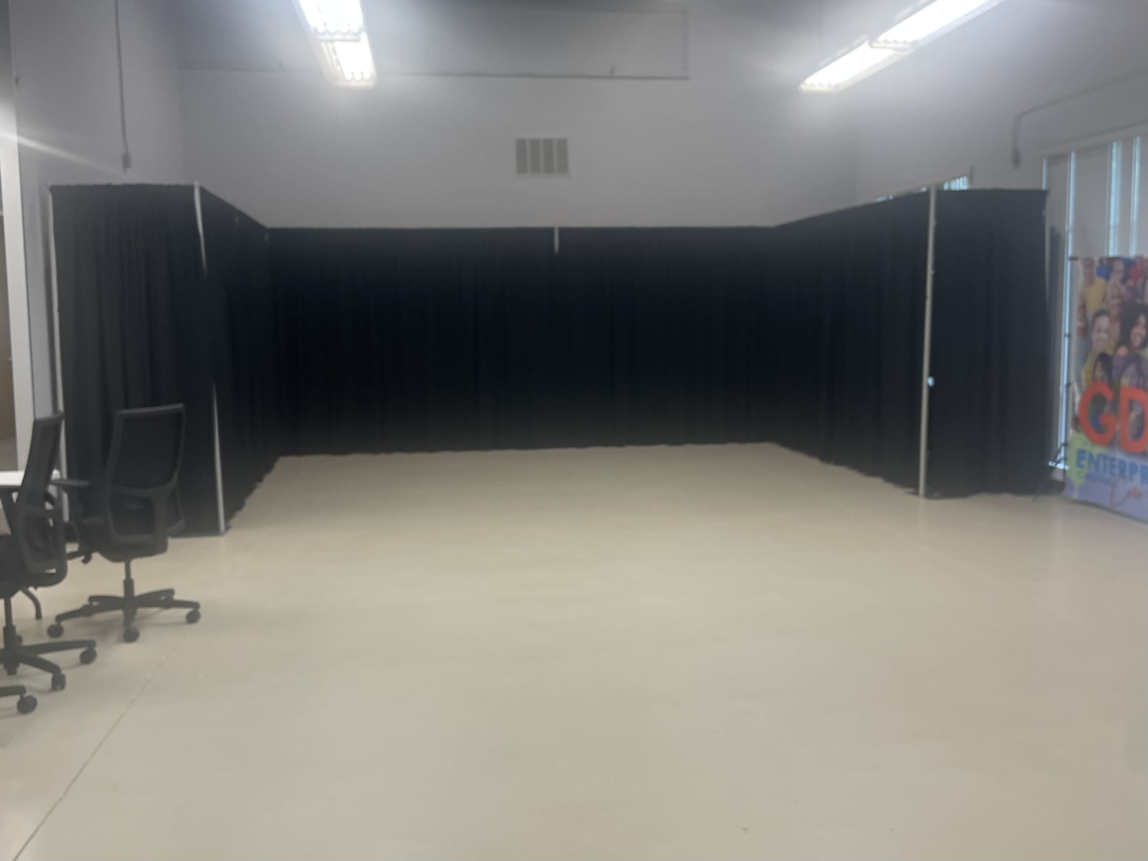 A large, empty room divided by black curtains, with a few chairs positioned nearby and a colorful banner in the corner.