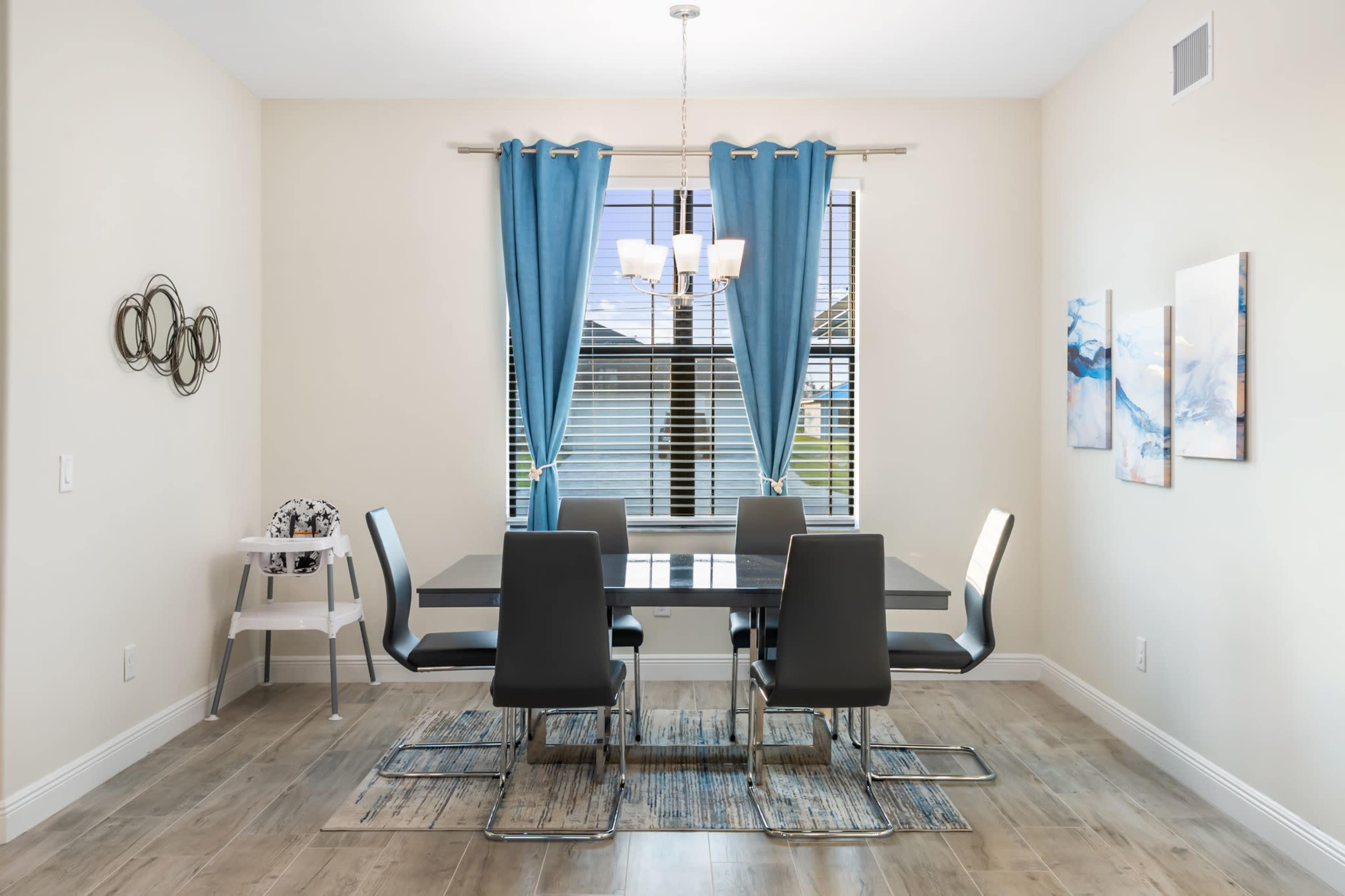 A modern dining room features a glass table surrounded by six black chairs, with blue curtains and artwork on the walls.