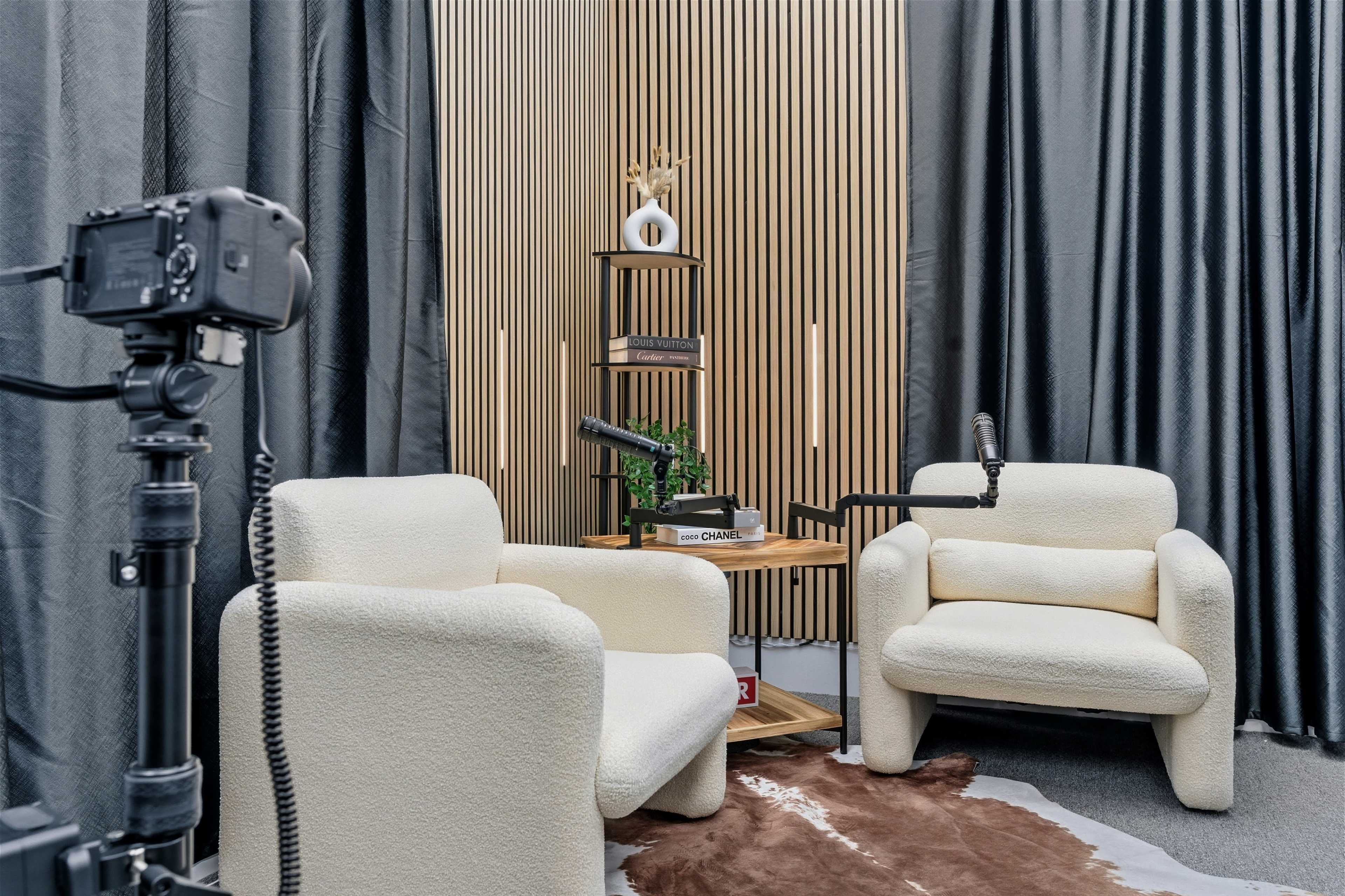 The image shows a cozy interview setup featuring two white armchairs, a wooden shelf with decorative items, and a microphone, all against a backdrop of dark curtains.