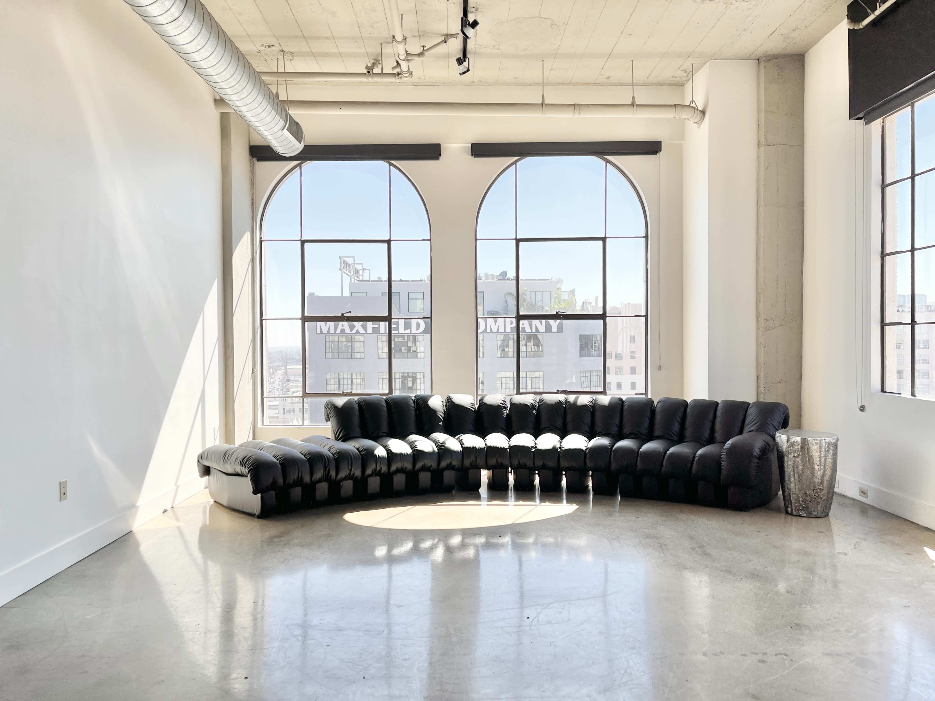 A black sectional sofa is positioned in front of large arched windows, casting long shadows on a polished concrete floor.