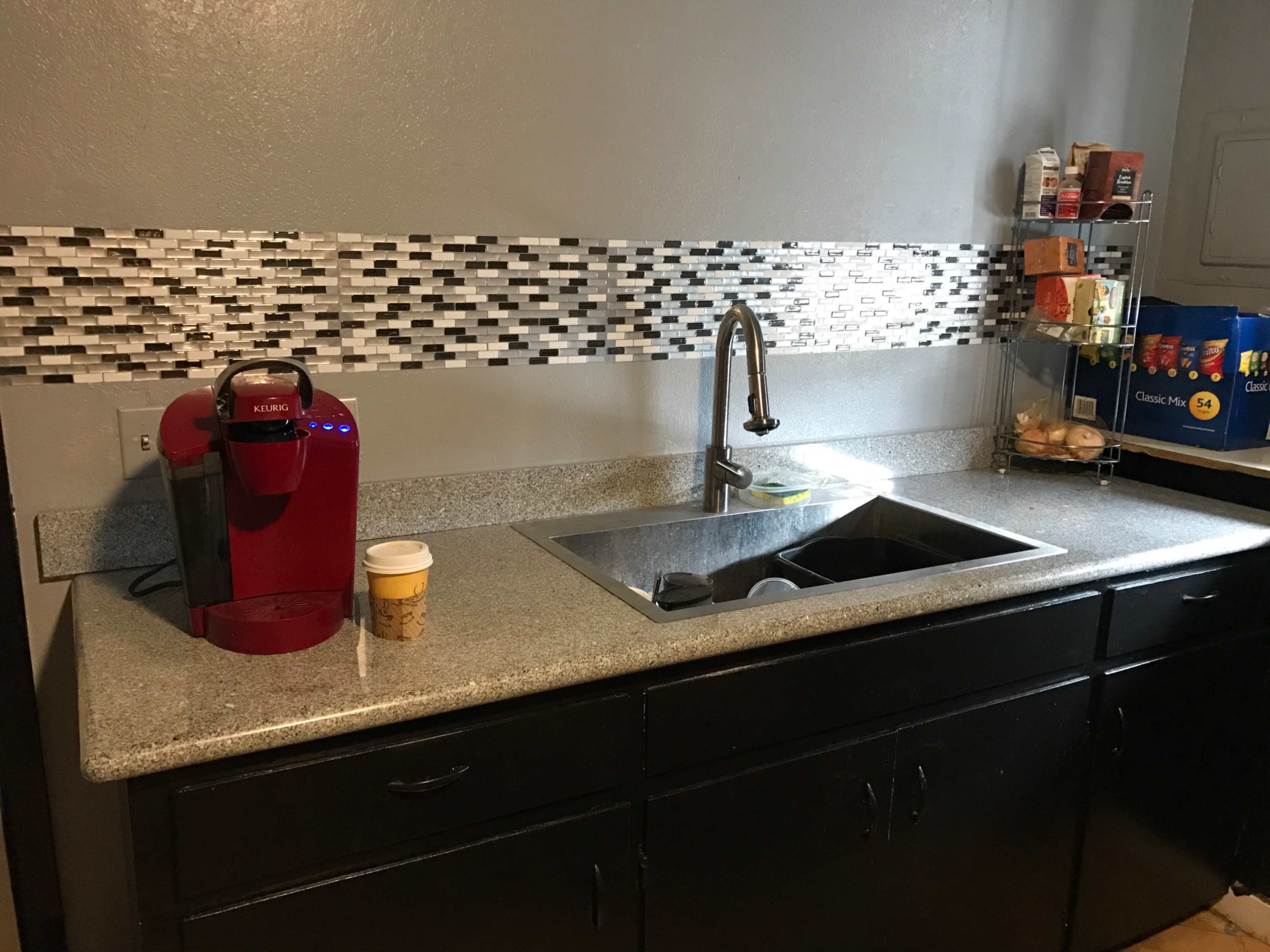 The image shows a kitchen countertop with a red coffee maker, a sink, and a mosaic backsplash, along with a variety of food items on shelves.