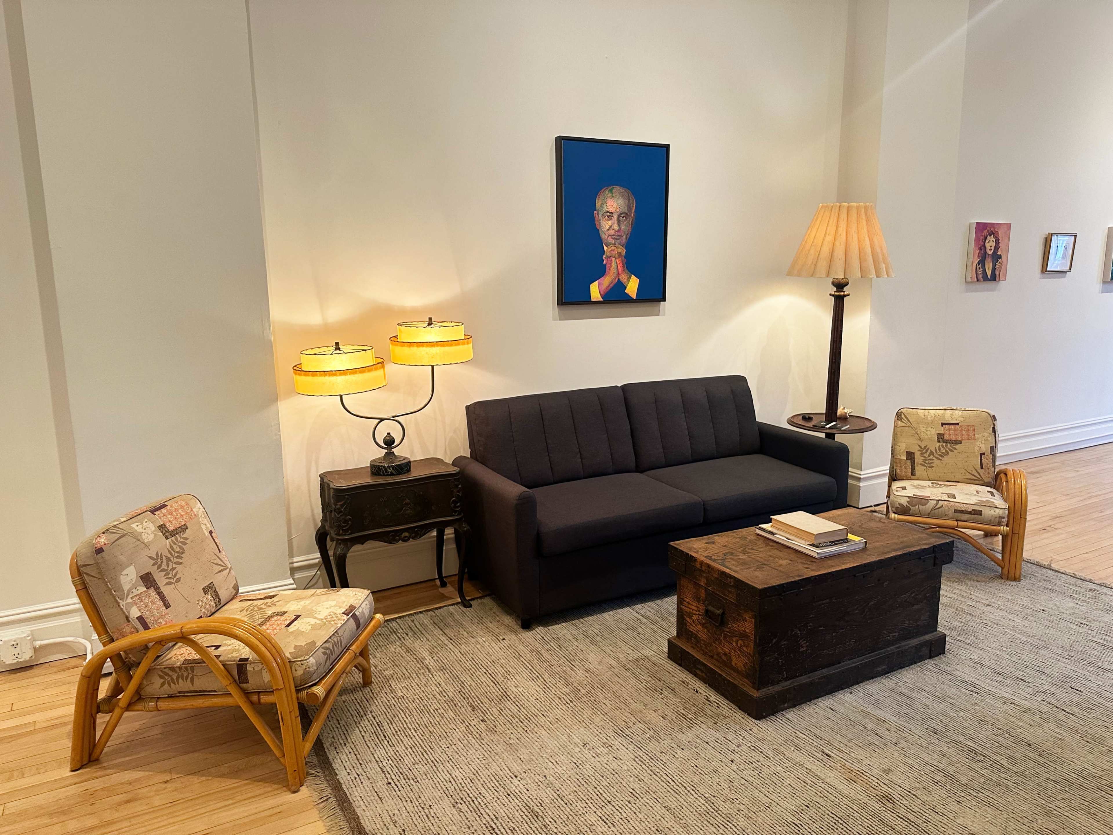 The image shows a minimalist living space featuring a dark gray sofa, two patterned armchairs, a wooden coffee table, and a lamp, with a painting of a person on the wall.