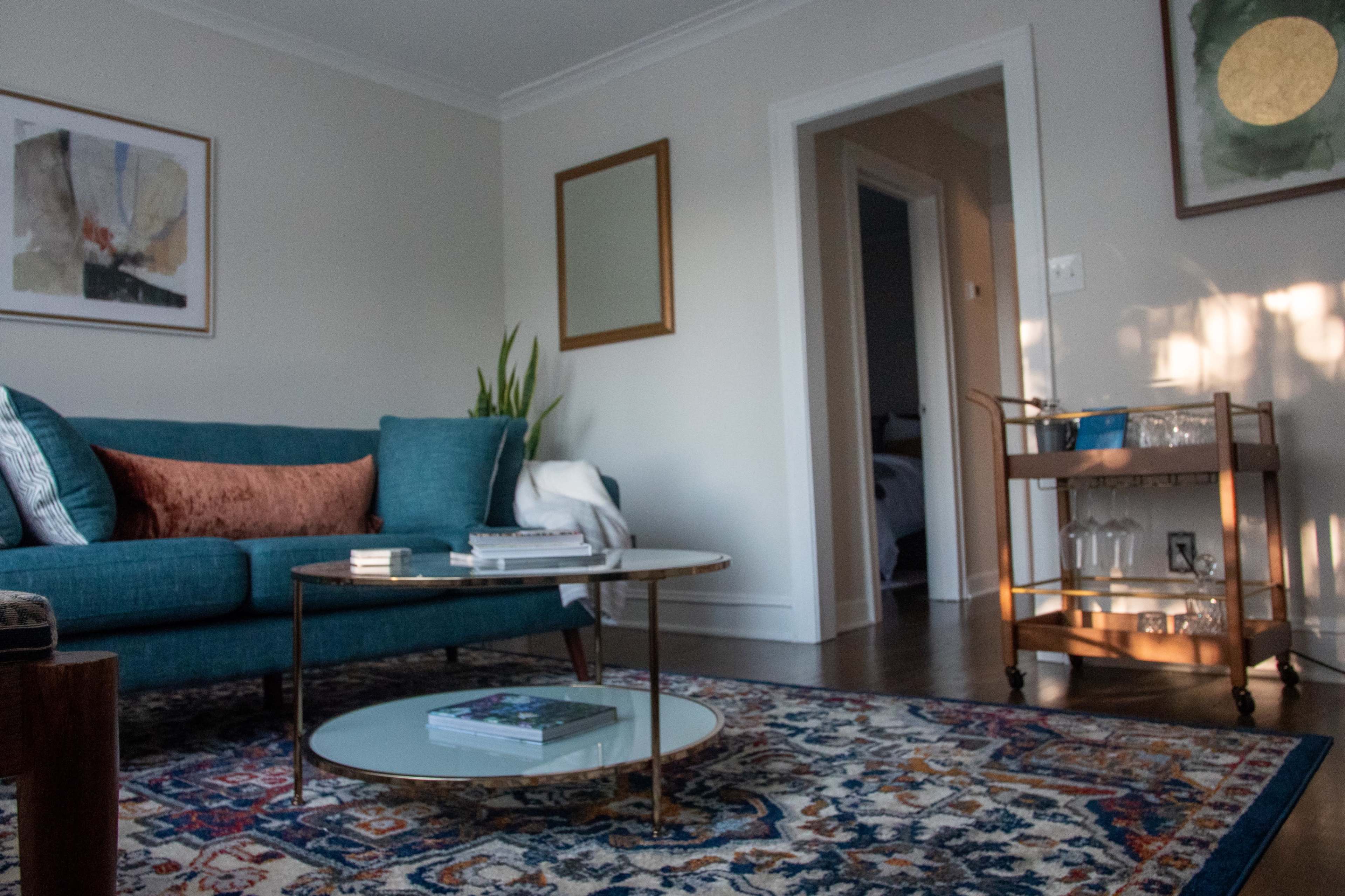 A living room features a teal sofa with decorative pillows, a glass-top coffee table, an ornate rug, and a small bar cart in the corner.