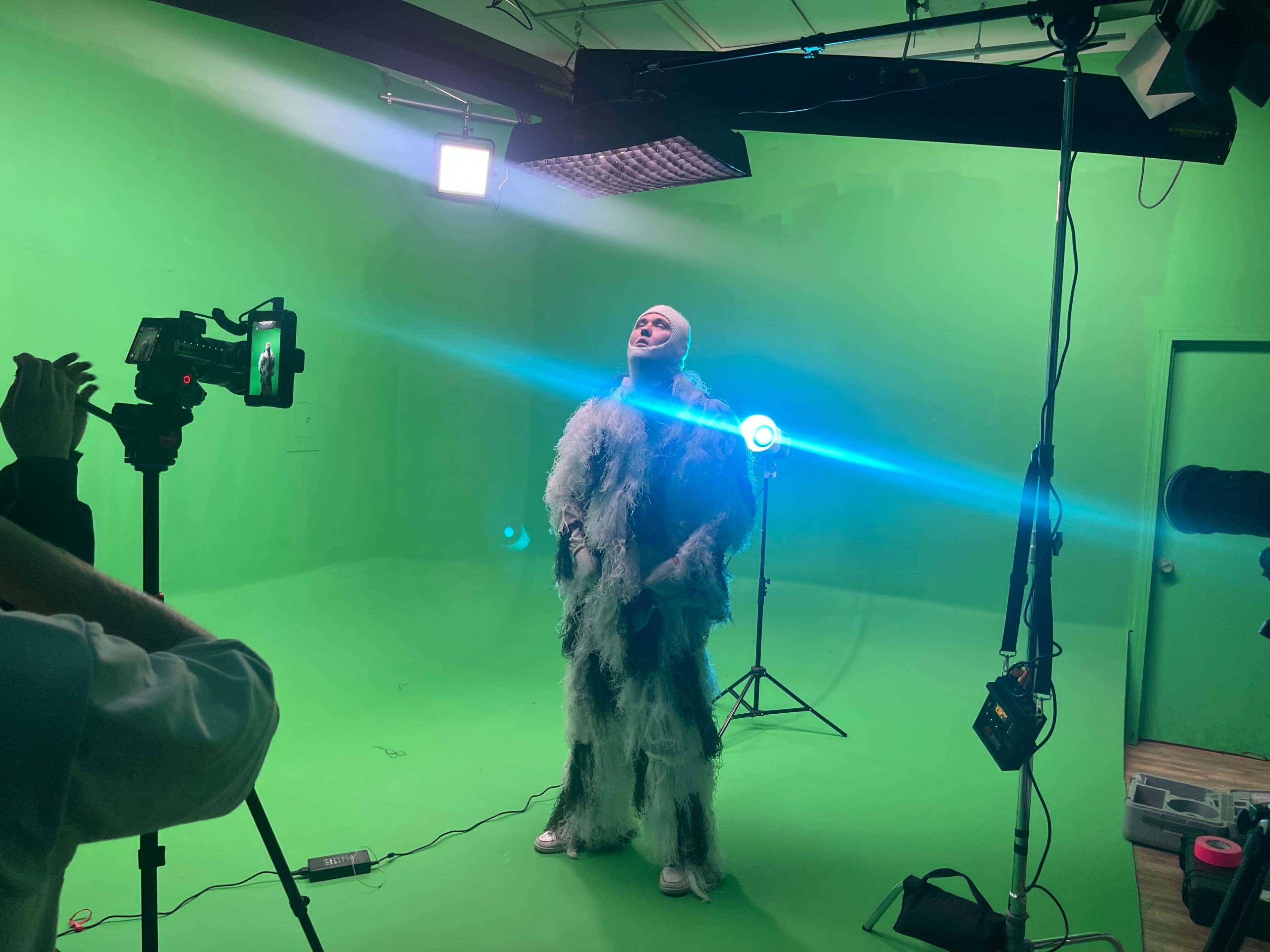 A person dressed in a fluffy costume stands against a green screen while being photographed by a camera crew.