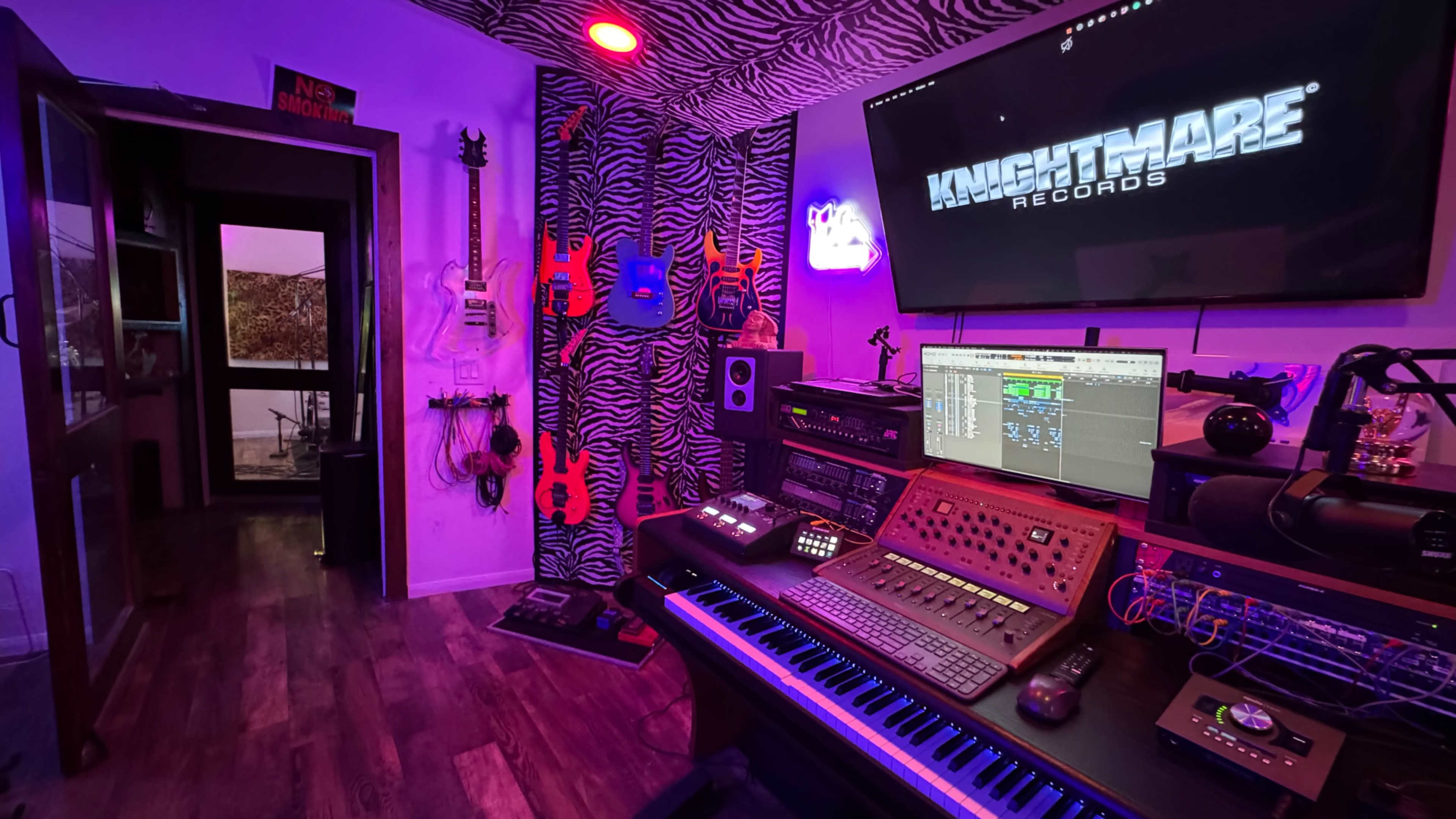 The image shows a music recording studio featuring a keyboard, audio equipment, vibrant lighting, and a wall adorned with various electric guitars.