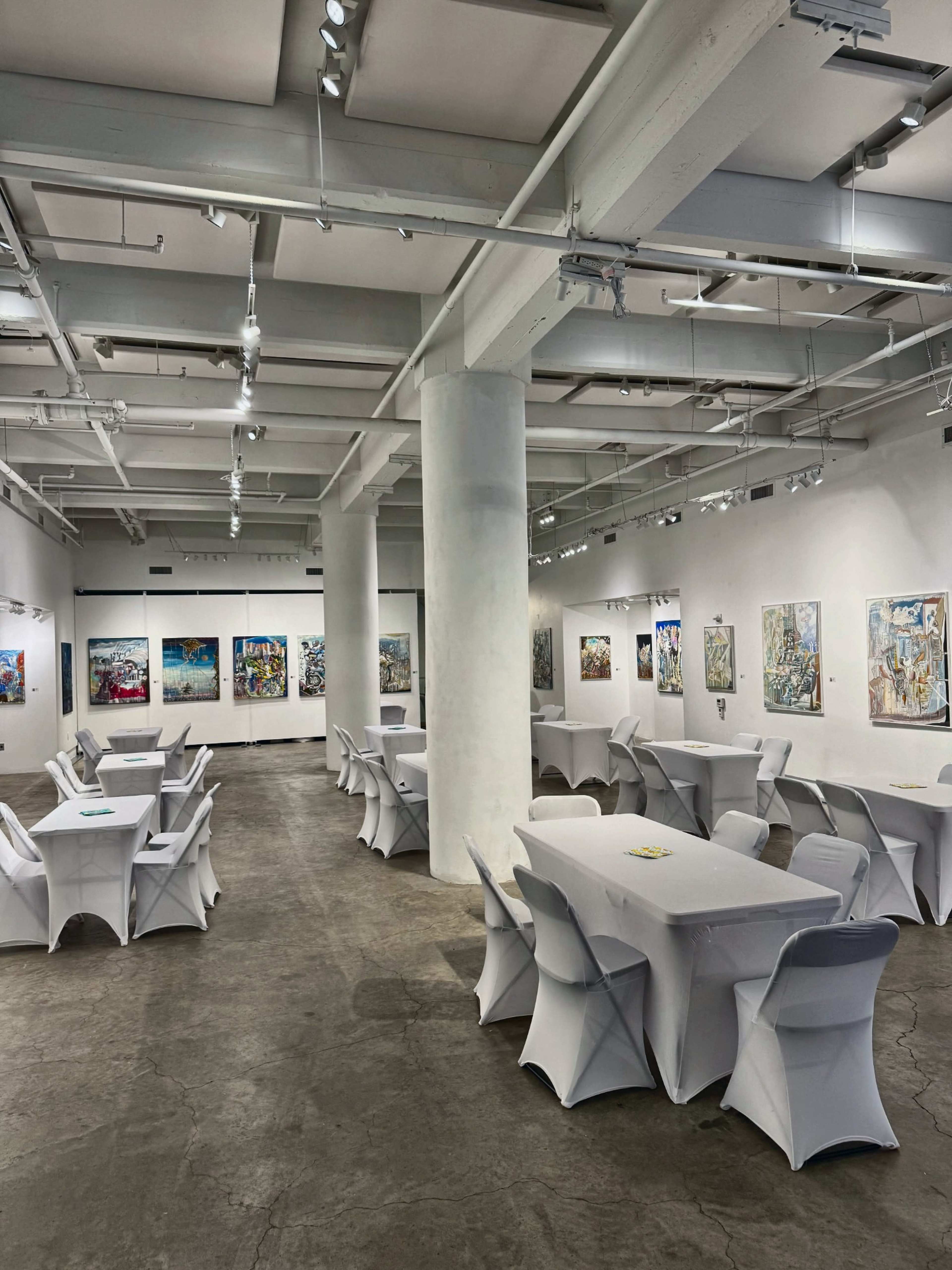 The image shows a spacious art gallery with white tables and chairs arranged in a symmetrical layout, surrounded by colorful paintings displayed on the walls.