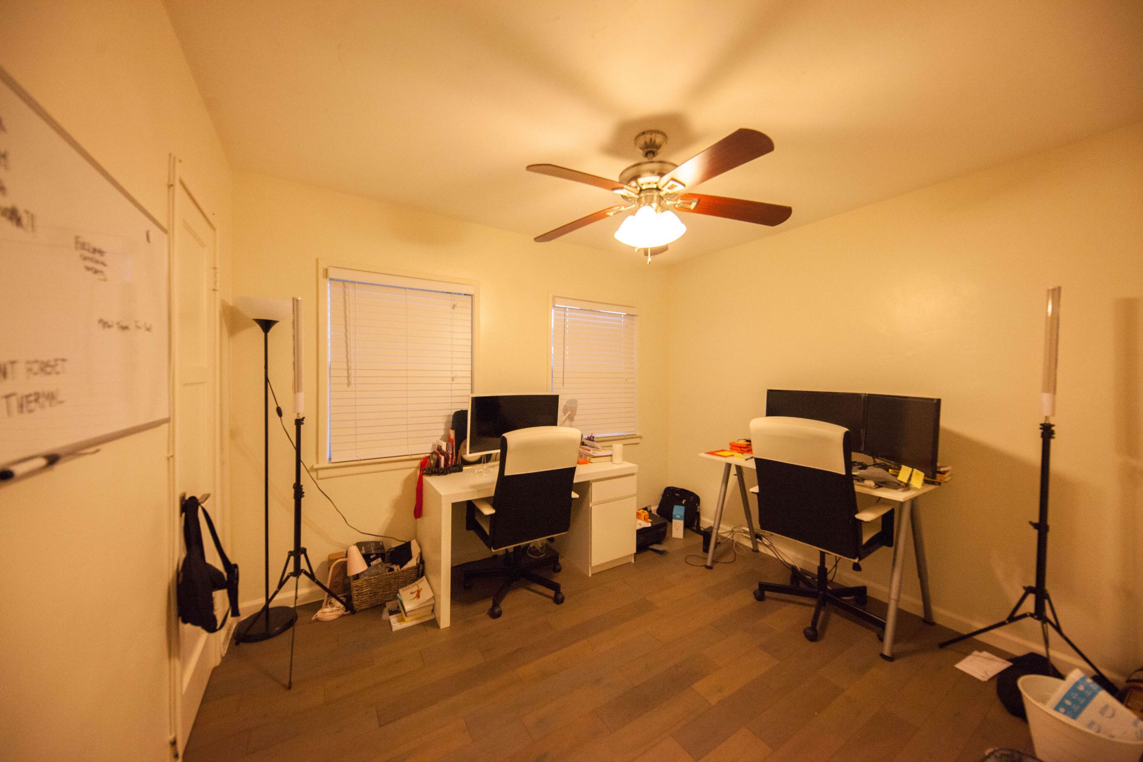 A small office space with two workstations, each featuring a computer and a chair, illuminated by a ceiling fan.