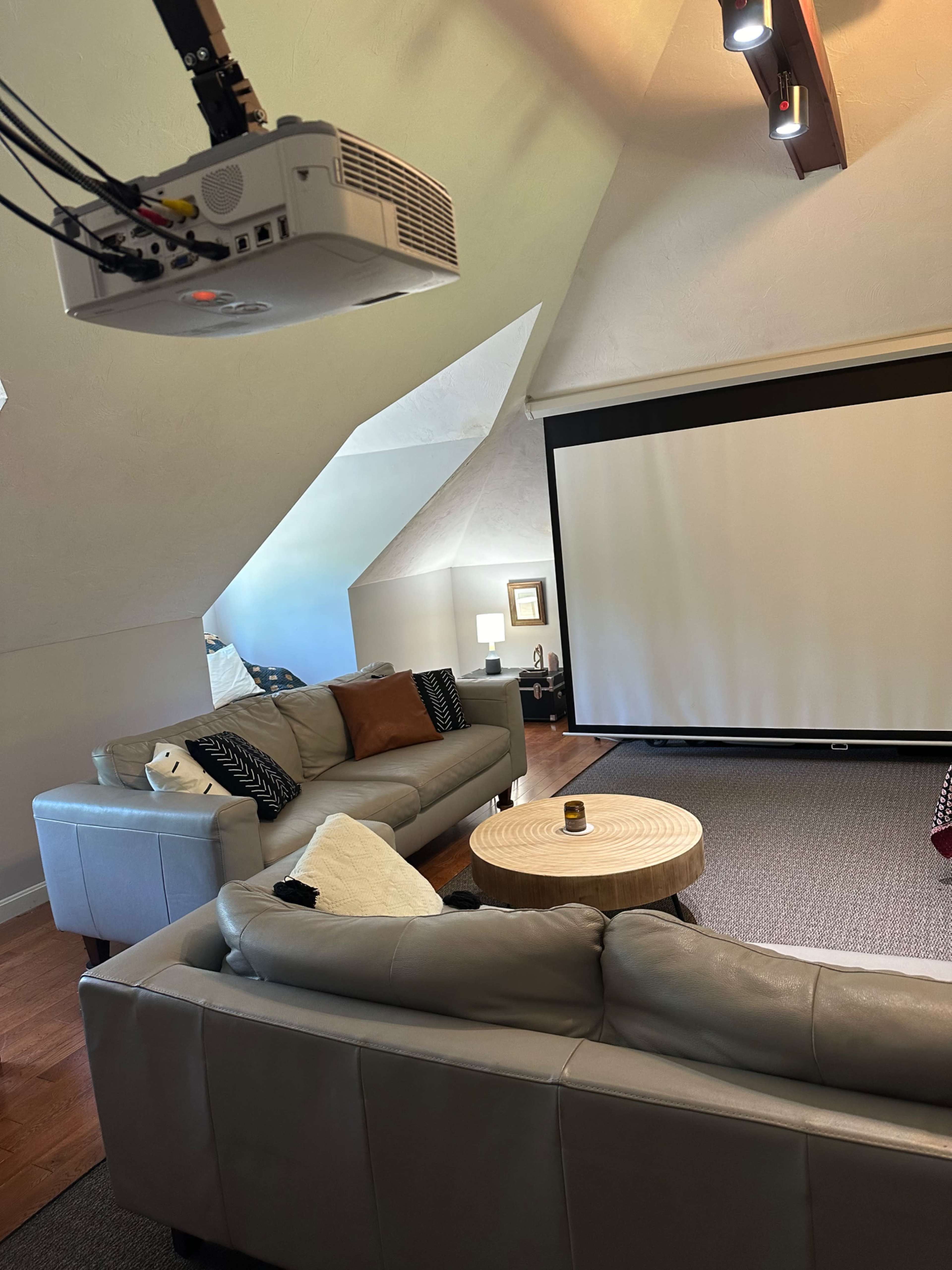 A spacious room with a projector mounted on the ceiling, two sofas facing a large projection screen, and a coffee table in the center.