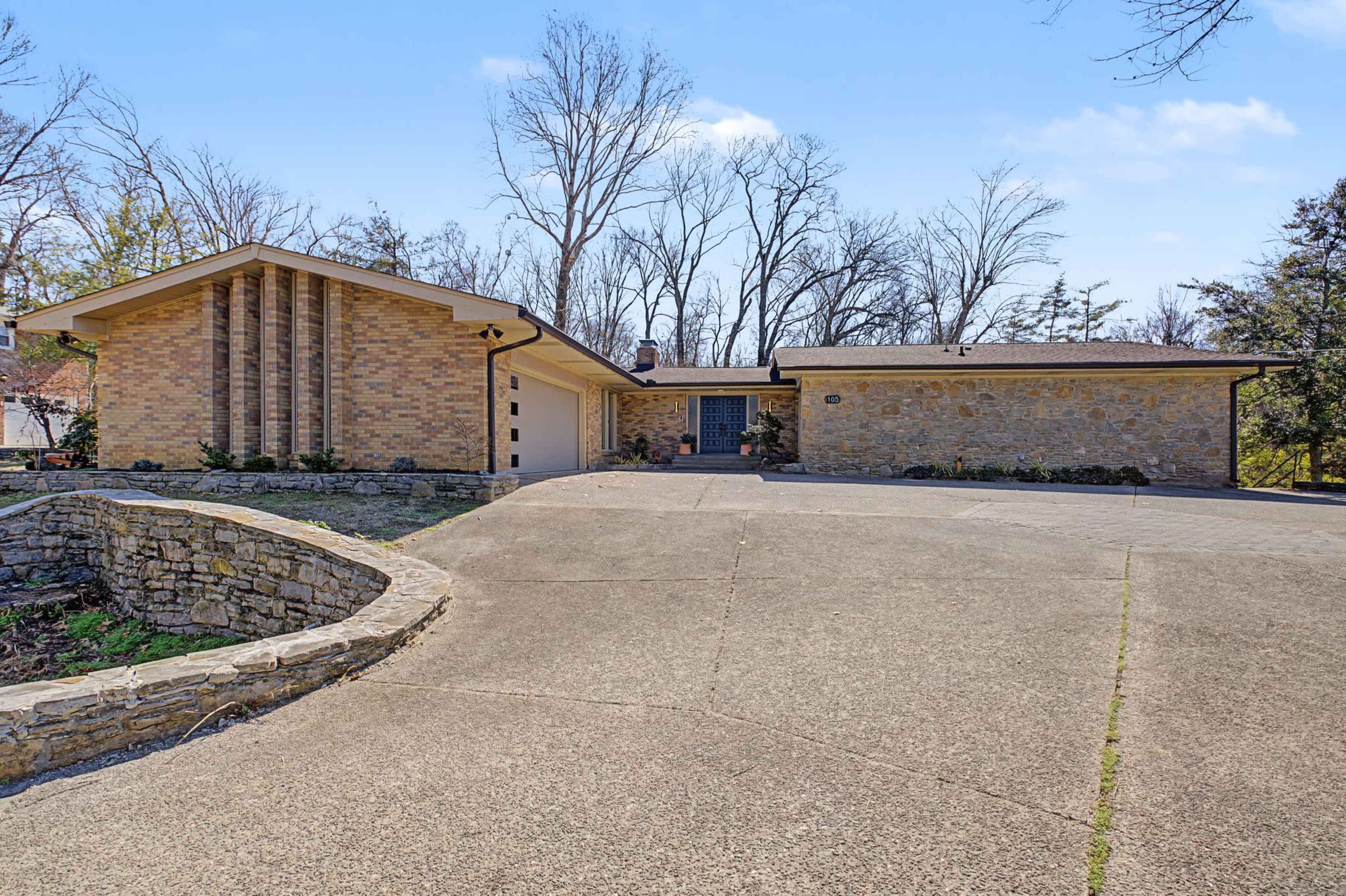The image depicts a single-story mid-century modern house with a stone facade surrounded by a paved driveway and trees in the background.