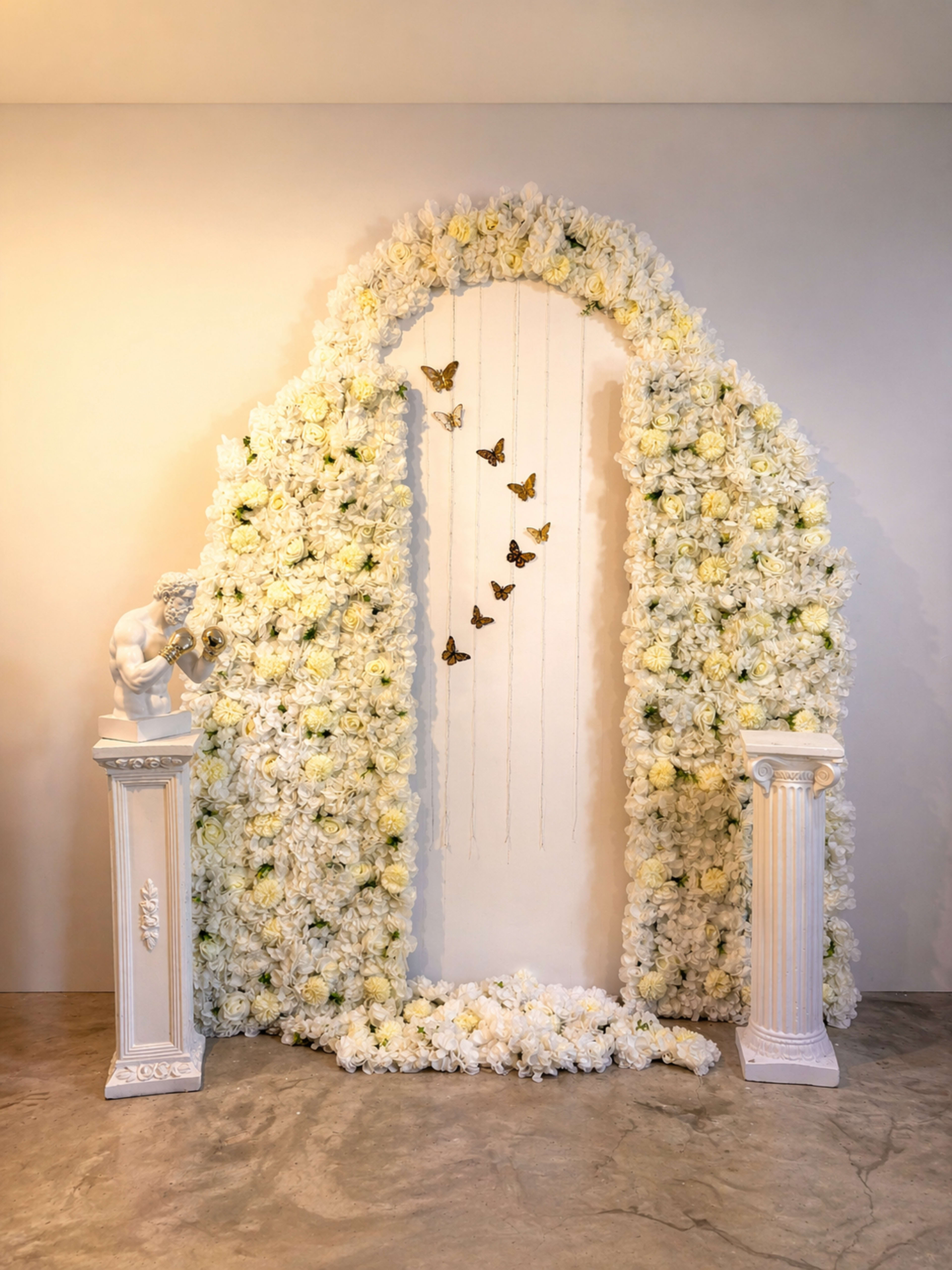 A decorative floral archway composed of white flowers is flanked by two white columns and features butterflies suspended from delicate strings.