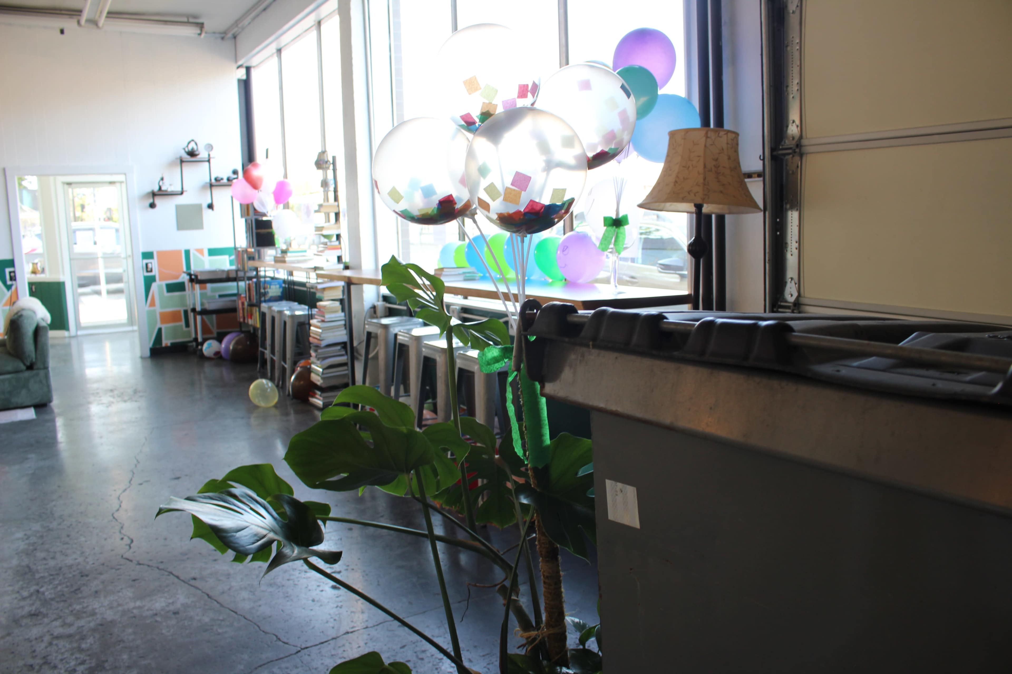 The image shows a room with large windows, colorful balloons, a potted plant, and a garbage bin in the foreground.