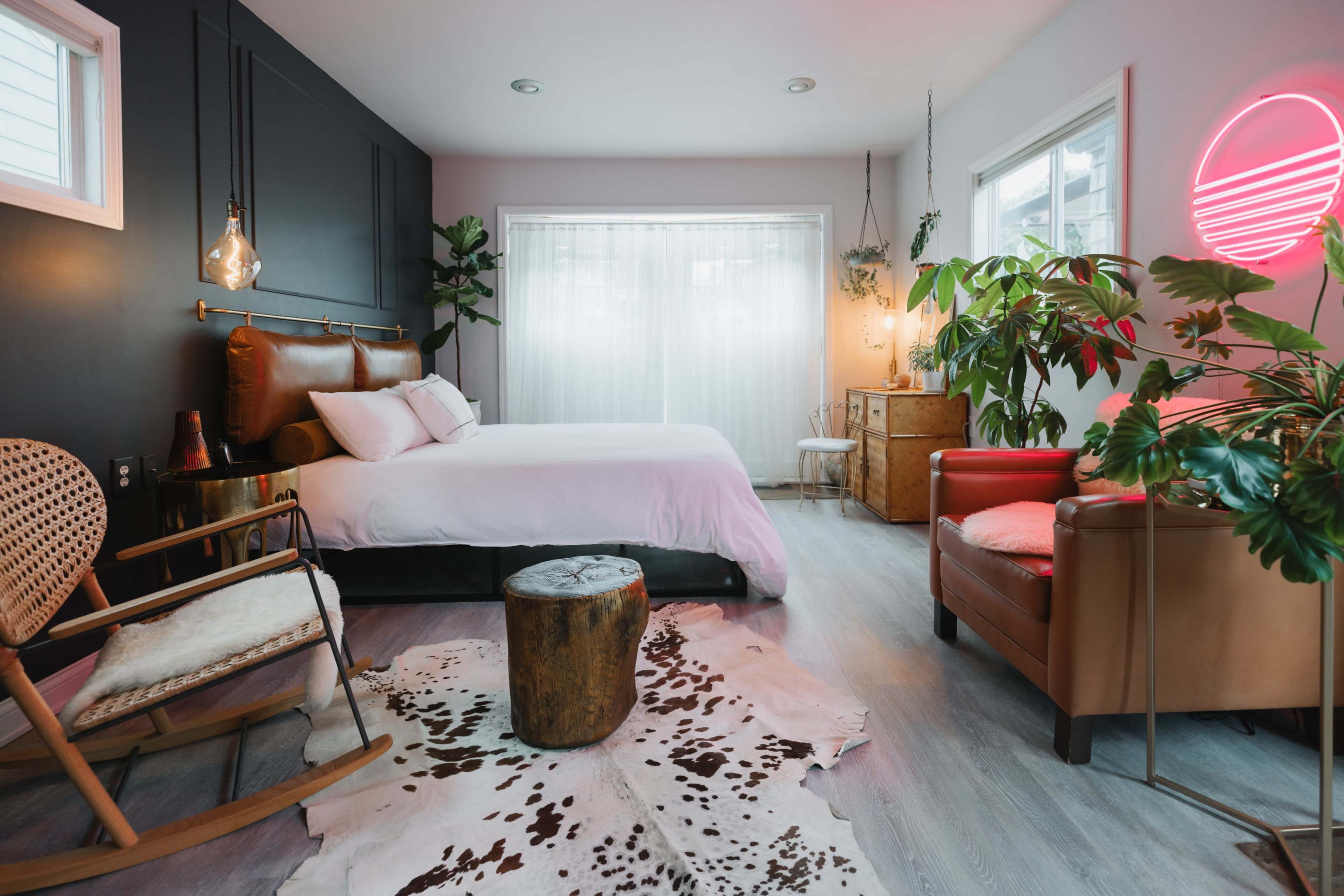 A modern bedroom featuring a bed with a brown leather headboard, a wooden rocking chair, a potted plant, and a neon light fixture.
