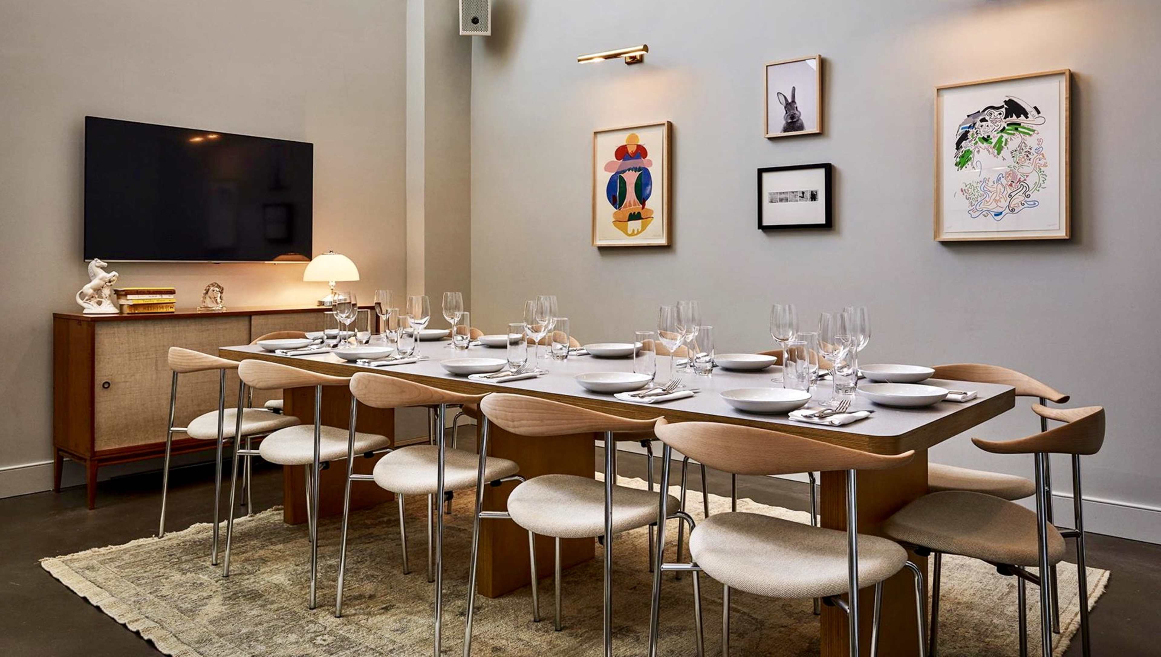 A modern dining room features a long table set for dinner, surrounded by chairs and decorated with framed artwork on the walls.