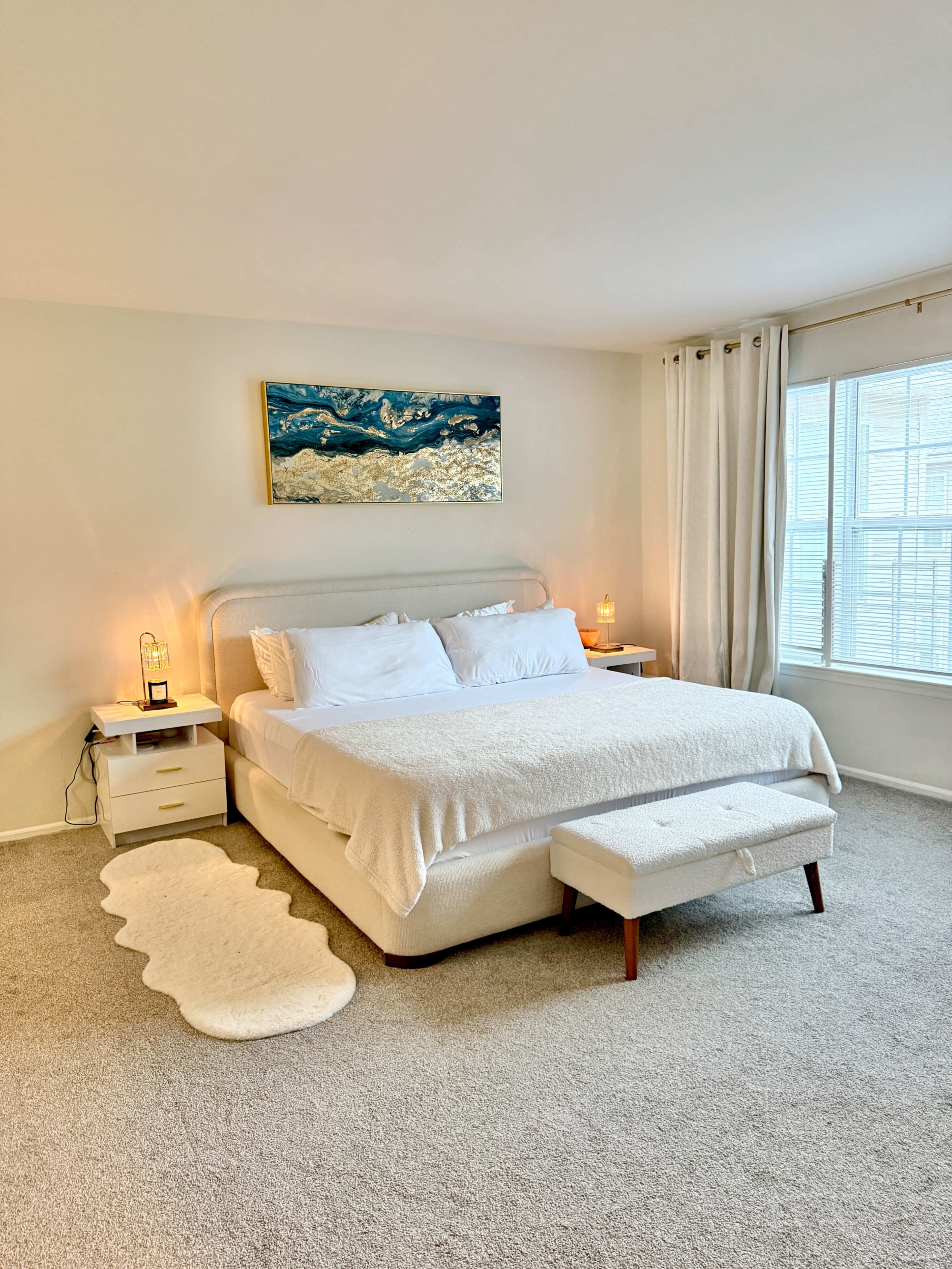 The image shows a neatly arranged bedroom with a large bed, two side tables with lamps, a bench at the foot of the bed, and a wall-mounted painting above the bed.