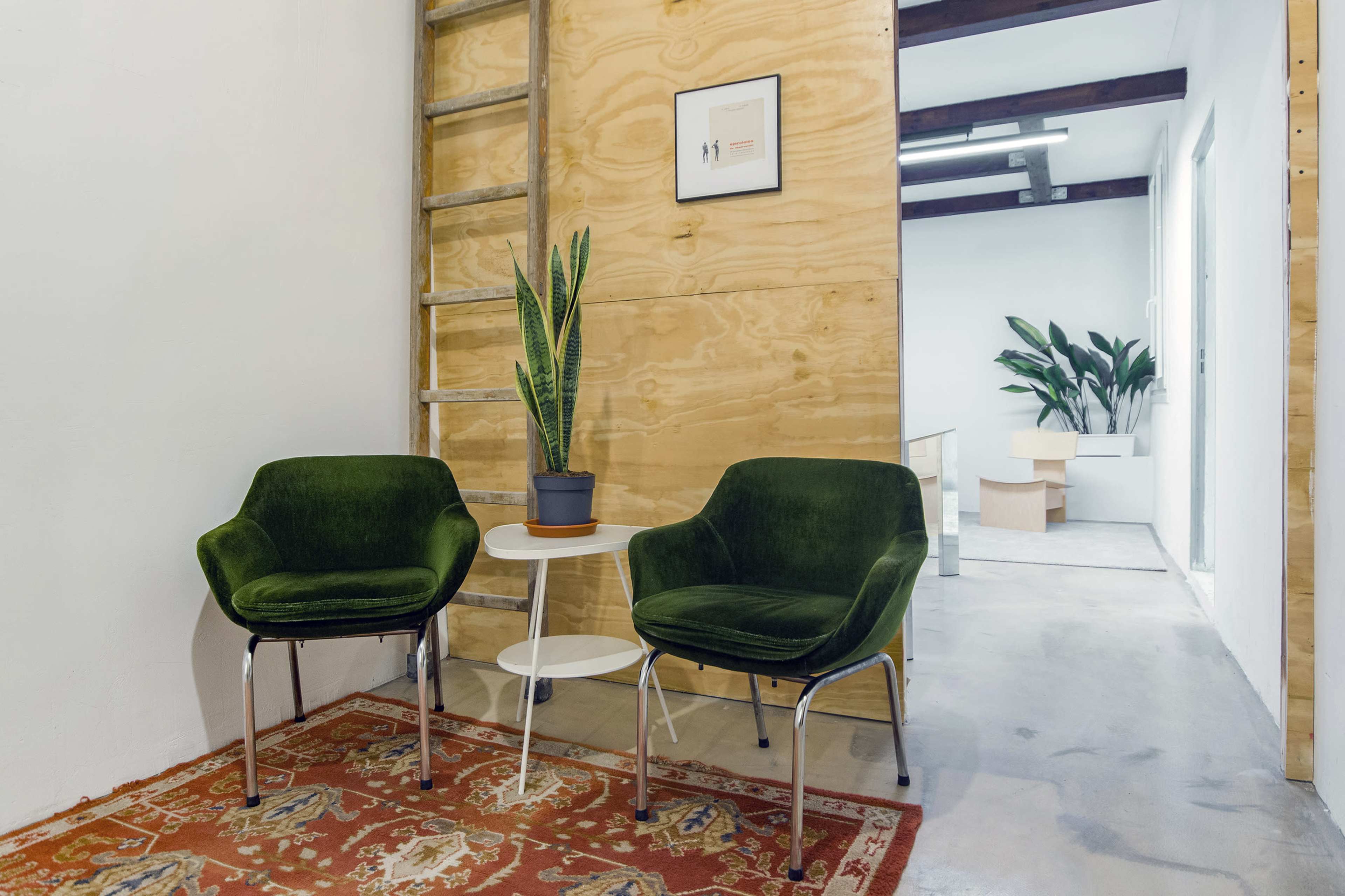 The image features two green chairs beside a small white table on a patterned rug, with a potted plant and a wooden ladder against a plywood wall.