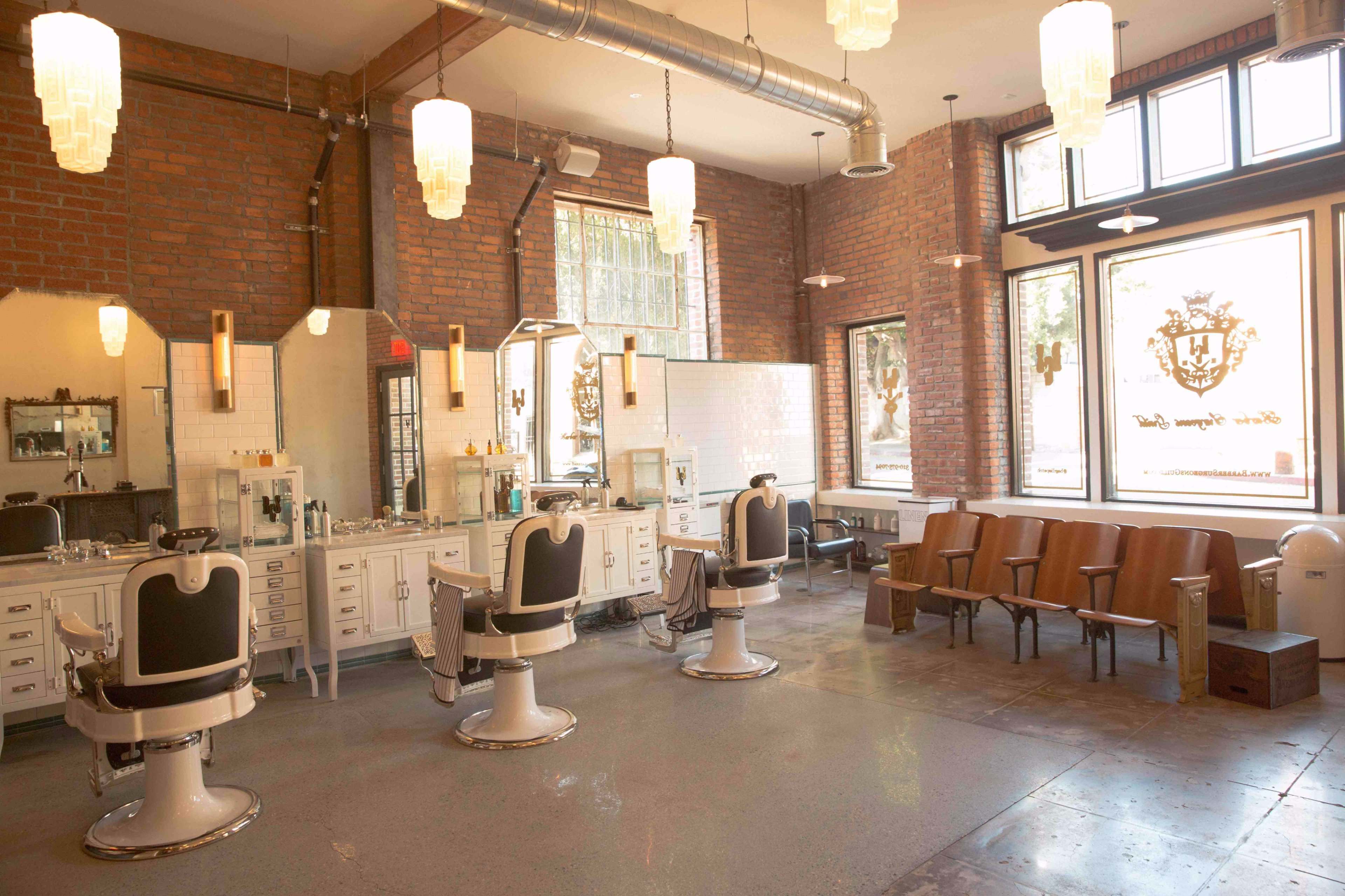 The image shows a modern barbershop with vintage-style barber chairs, large mirrors, and wooden seating against a backdrop of exposed brick walls.