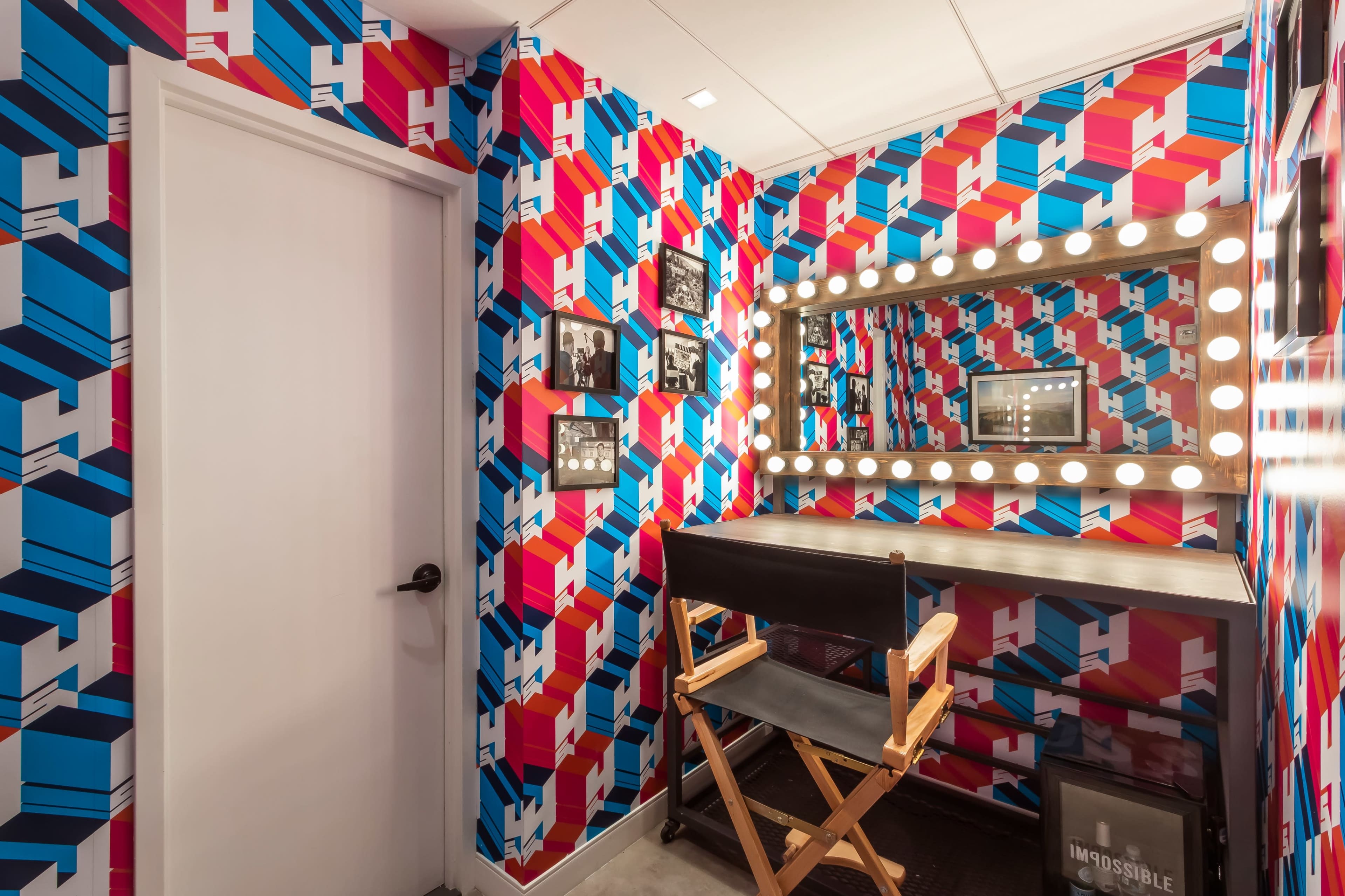 A brightly colored dressing room with a patterned wall, a large illuminated mirror, and a director's chair.
