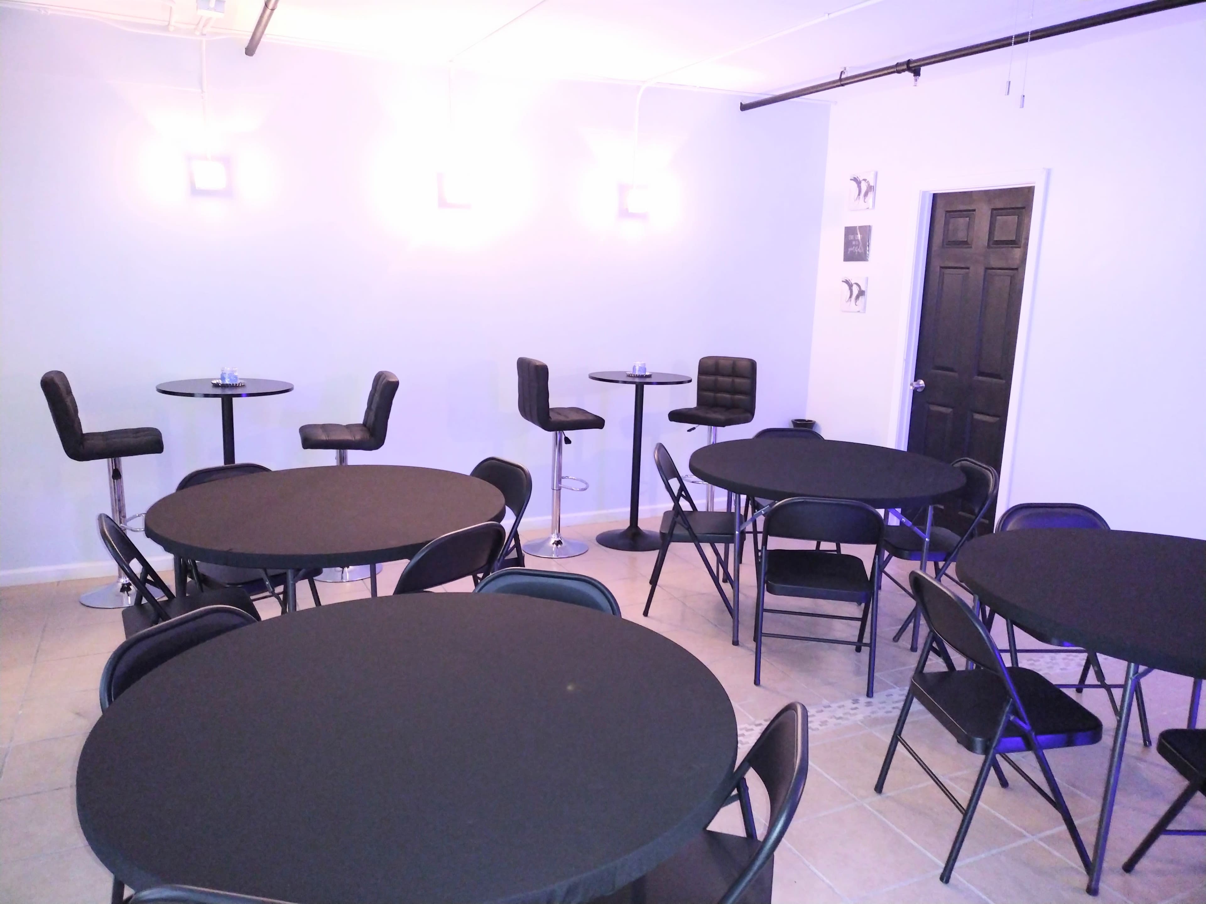 The room features several round tables with black tablecloths, high-top stools, and a black door in one corner.