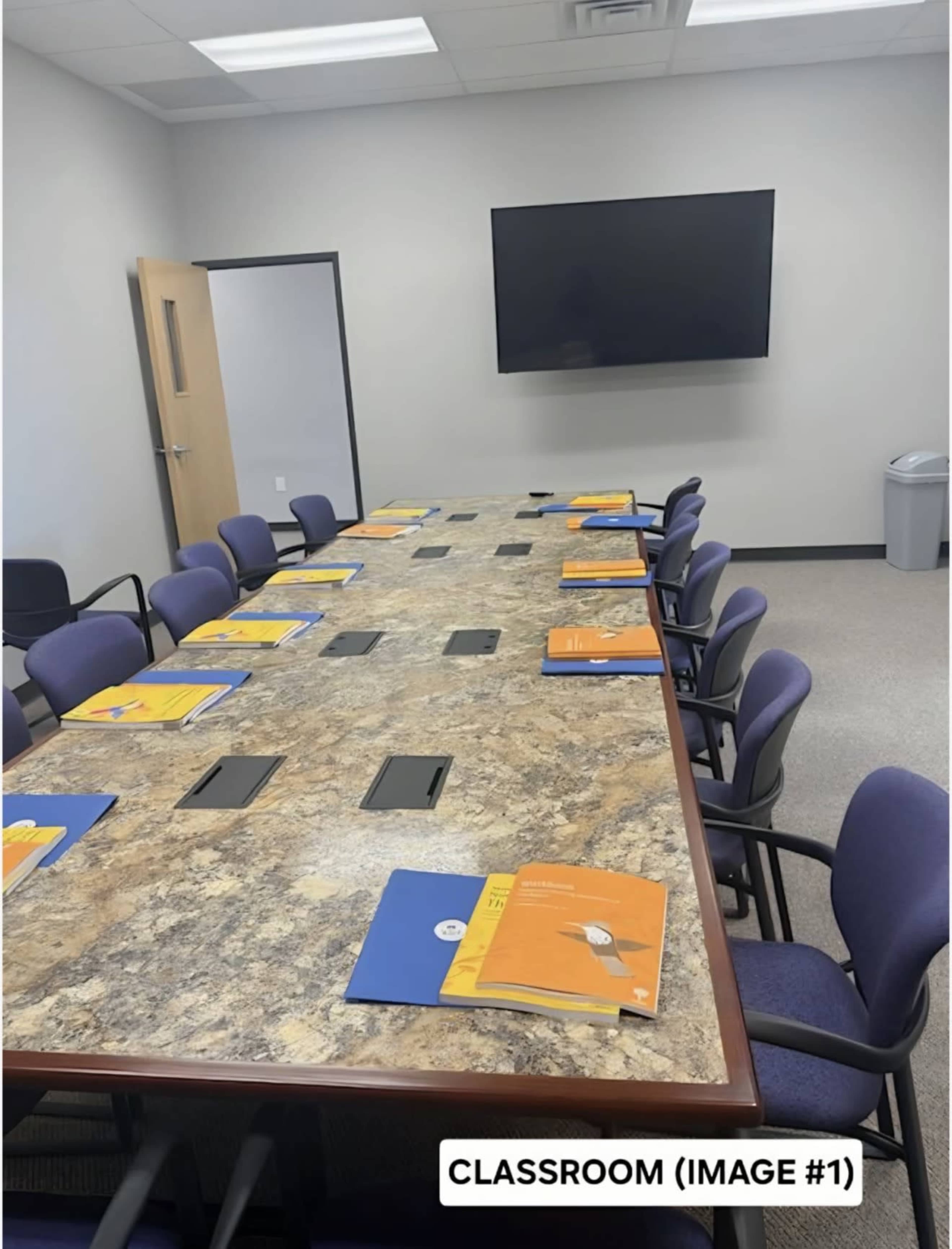 A long conference table is set with folders and notebooks, and a large screen is mounted on the wall in a modern classroom.