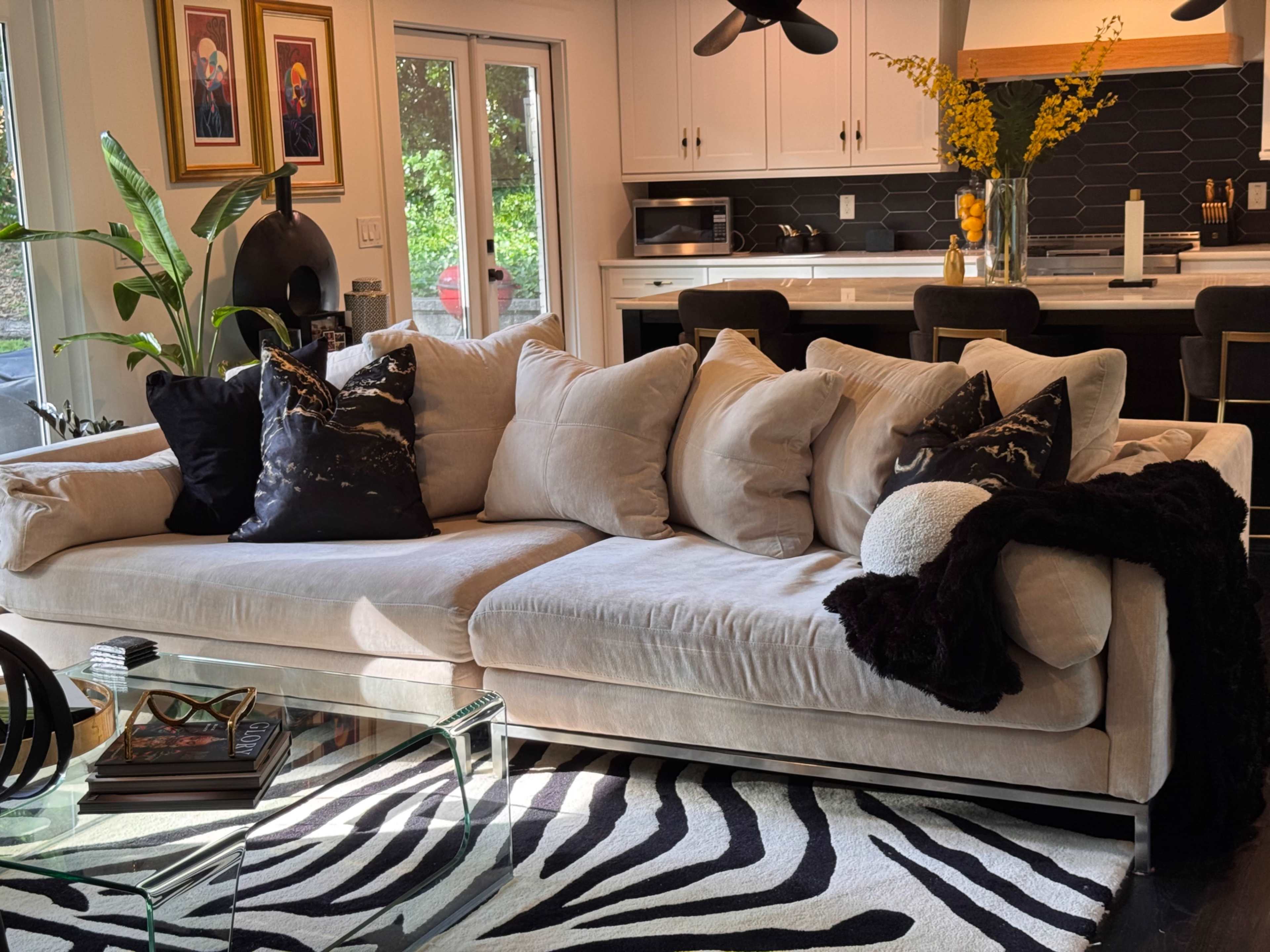 The image features a spacious living area with a large beige sectional couch adorned with black and white decorative pillows, a glass coffee table, and a zebra-print rug, all set against a backdrop of a modern kitchen.