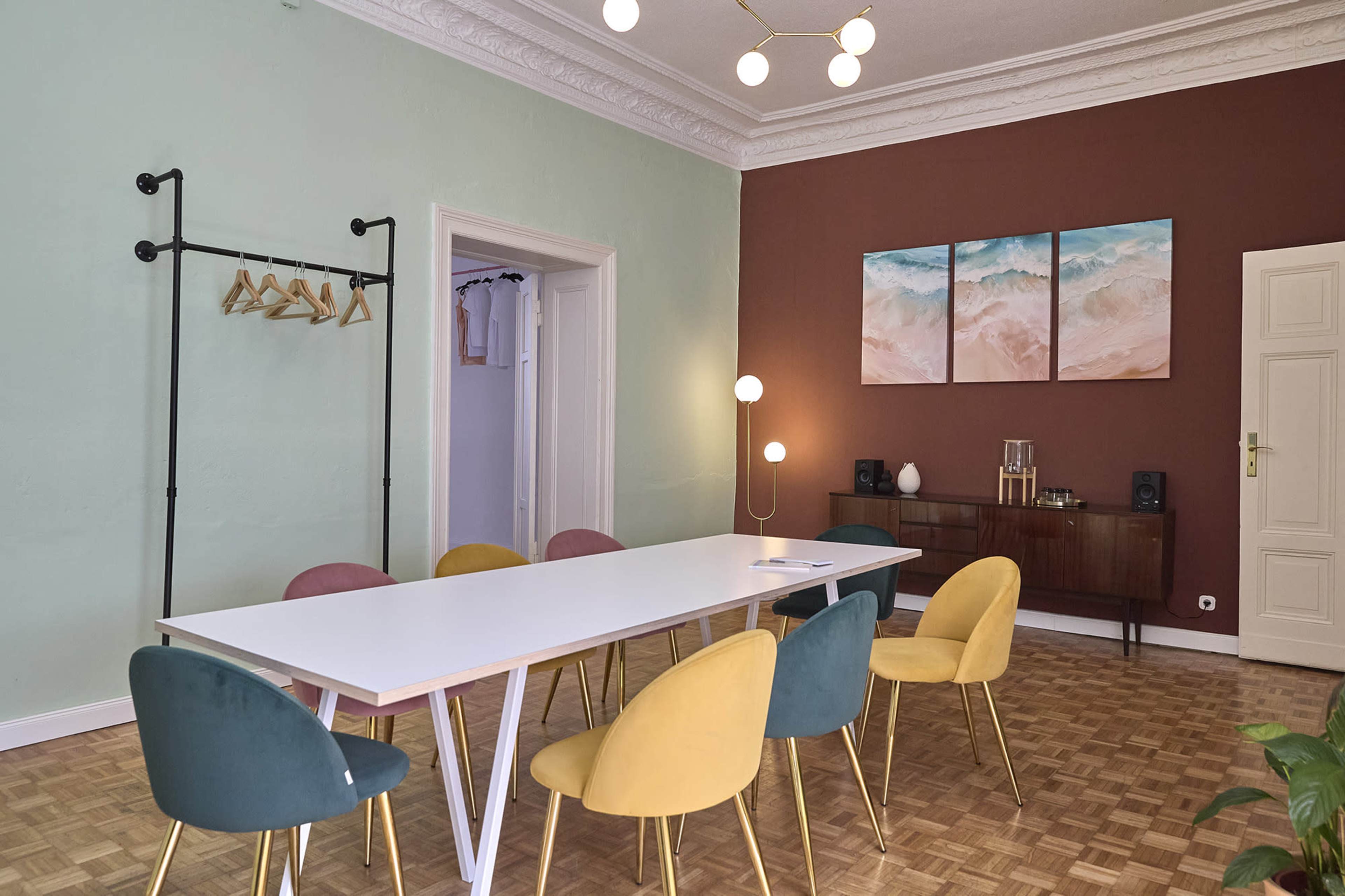 The image features a modern dining area with a long white table surrounded by colorful chairs, and abstract artwork on the wall.
