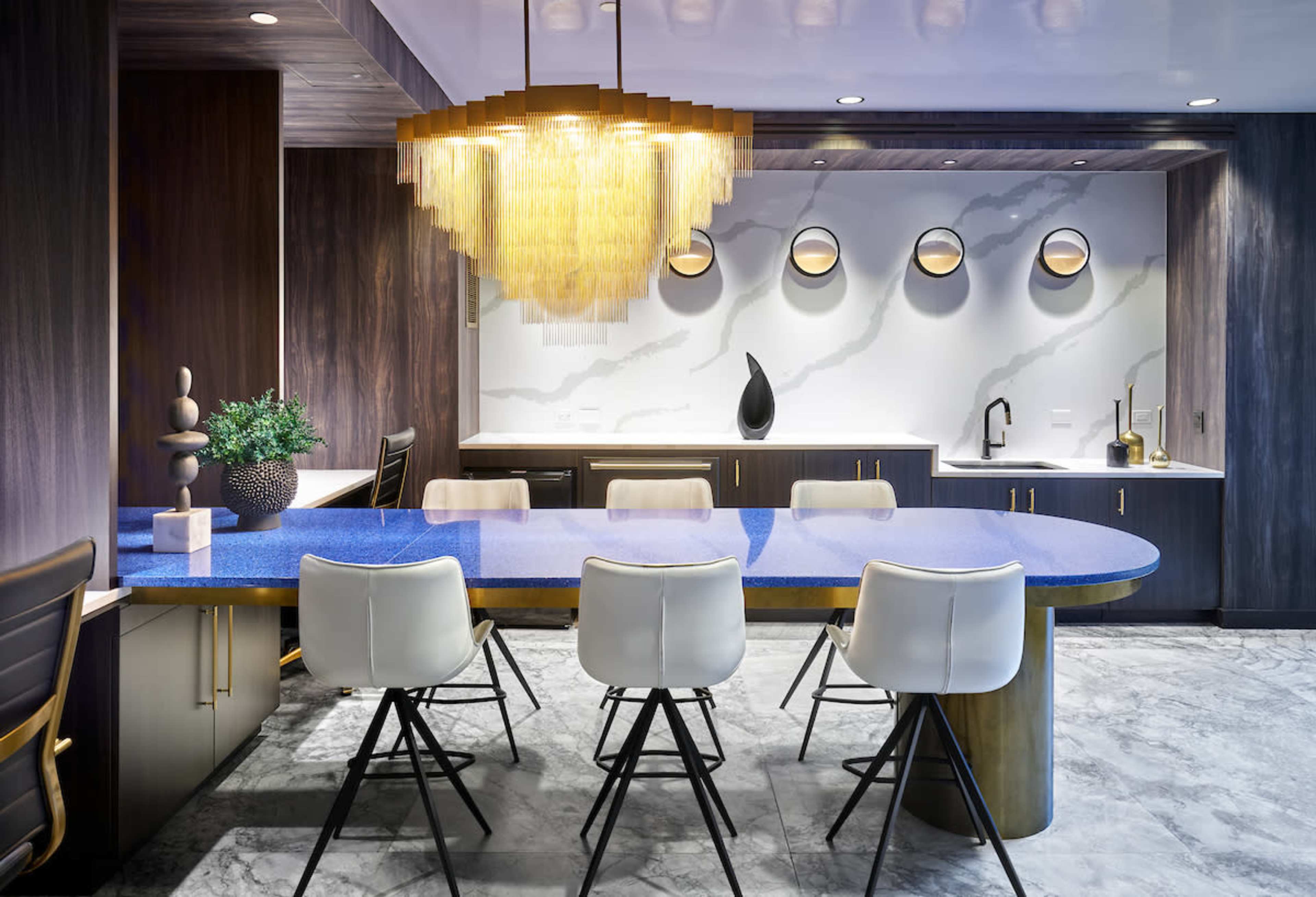 The image shows a modern dining area with a blue granite table, white chairs, and a chandelier above, set against a wall with decorative round mirrors and dark wood cabinetry.